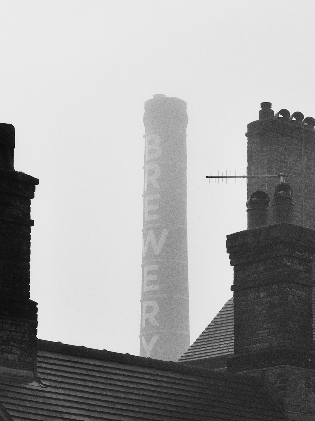 A black and white photograph of a tall smokestack with the word "BREWERY" written vertically on it. The smokestack is partially obscured by the roof of a building in the foreground. The sky is overcast, creating a moody and somewhat somber atmosphere. The image conveys a sense of industrial history and the passage of time.