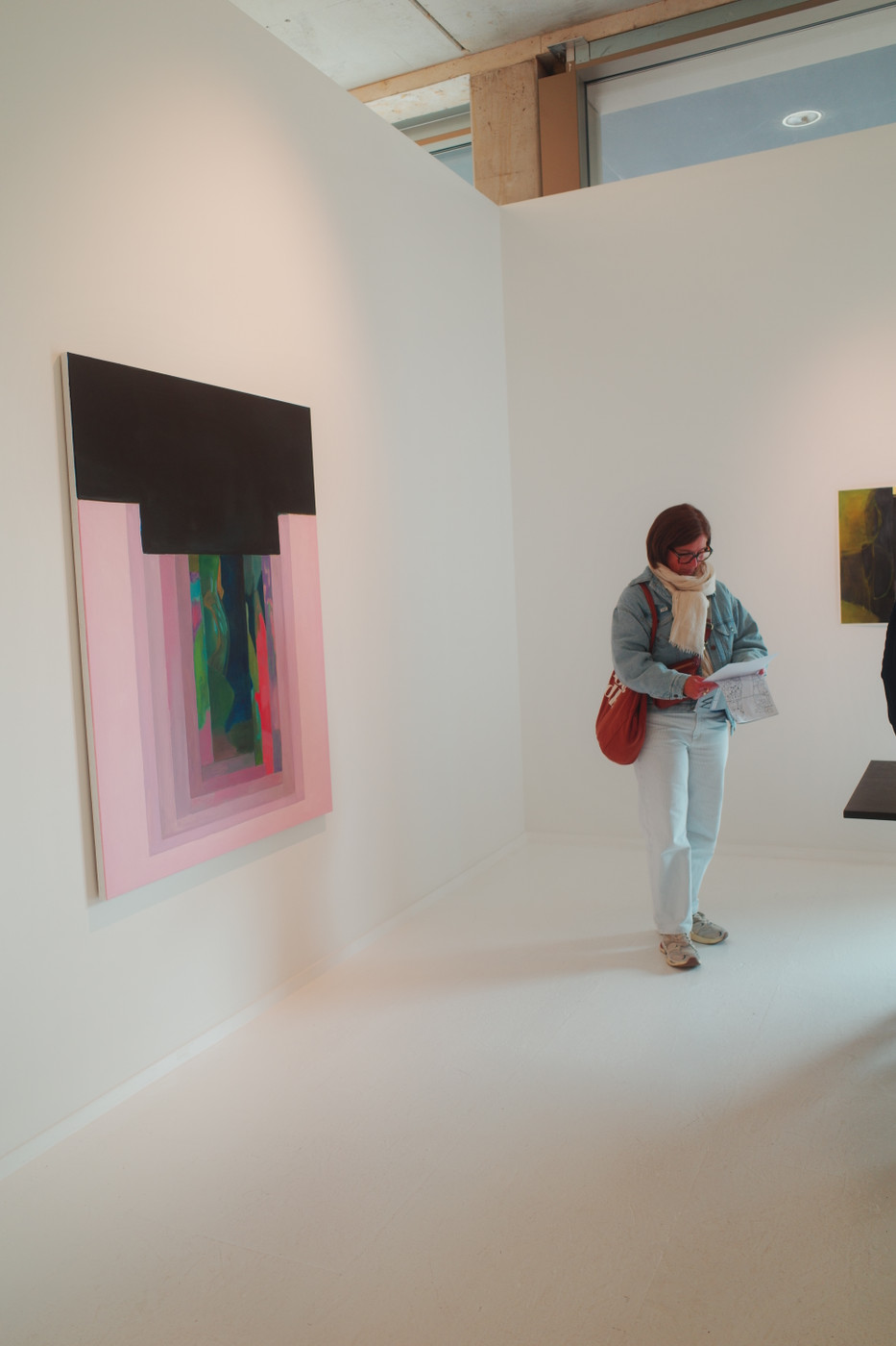 A spacious art gallery with white walls. A woman in a light scarf and denim jacket stands near a large abstract painting featuring black, pink, and multicolored elements, holding a folded paper and carrying an orange bag over her shoulder. A second painting is visible on the adjacent wall.