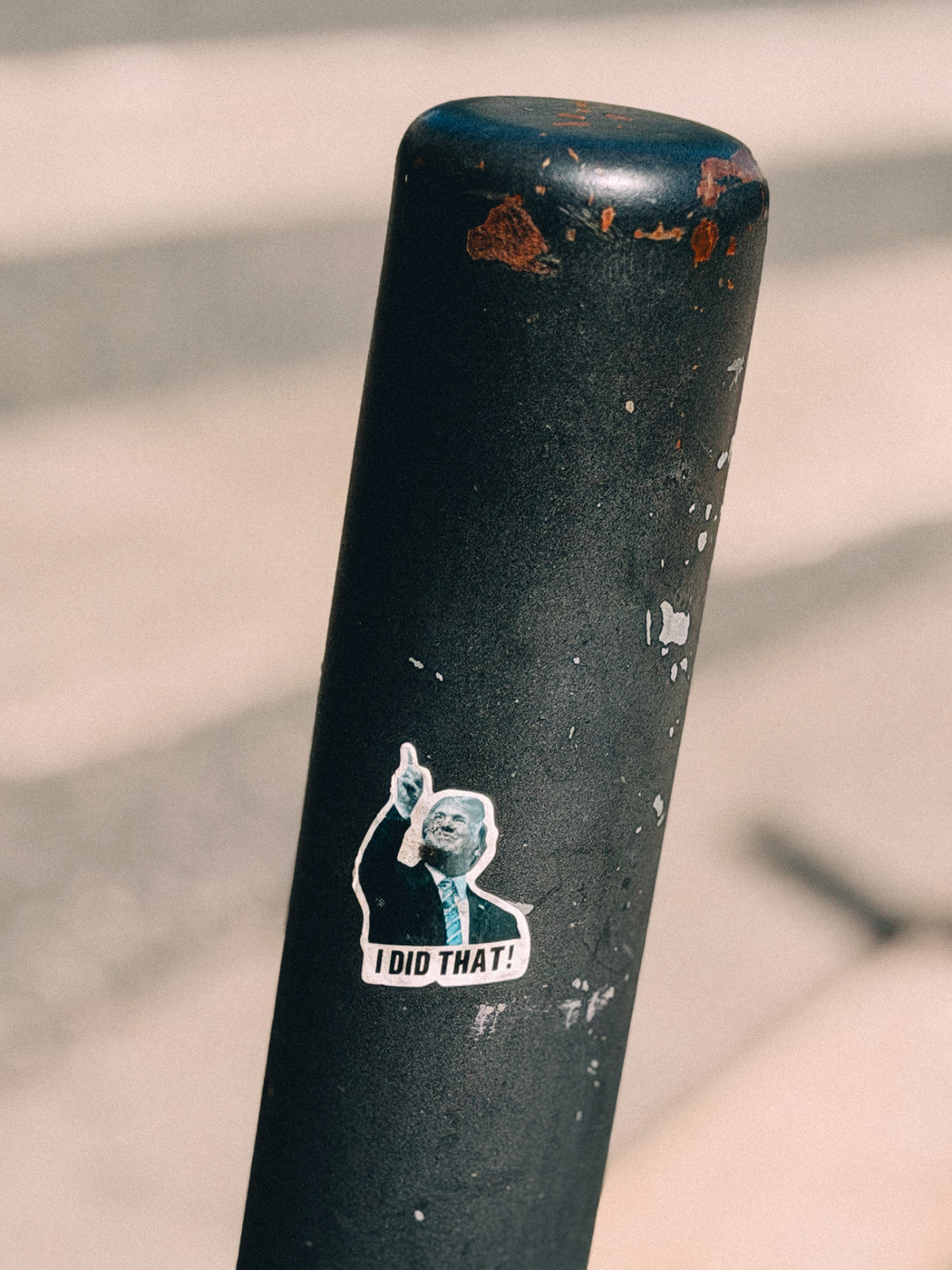 A weathered black pole with a small sticker of a man in a suit pointing upward, with the caption “I DID THAT!” on a blurred street background.