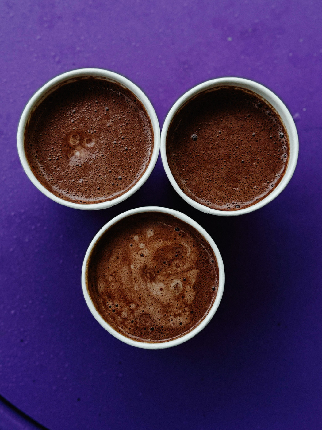 Three cups of hot chocolate on a purple surface arranged in a triangular pattern.