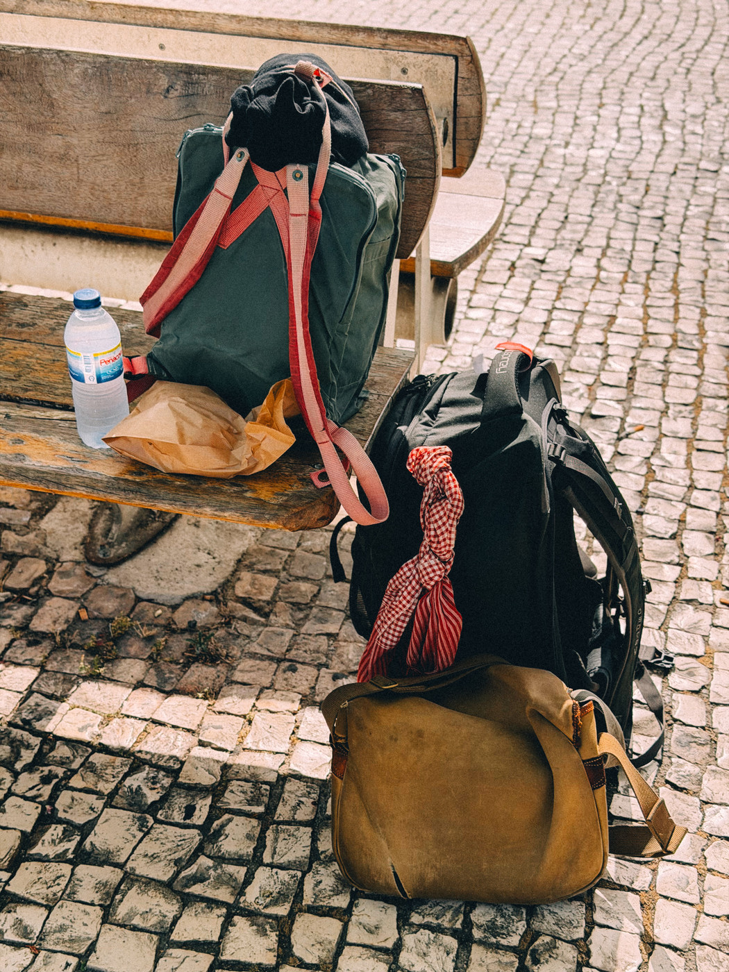 Three backpacks and a brown paper bag with a water bottle are on and beside a wooden bench on a cobblestone street.