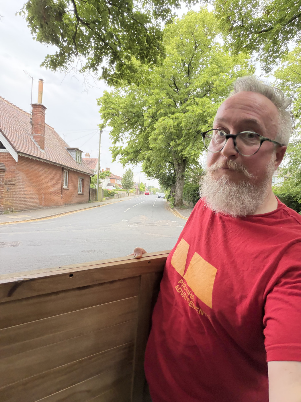 Man with grey beard and round glasses in a red graphic tee beside a large wooden furniture panel on a quiet residential street.