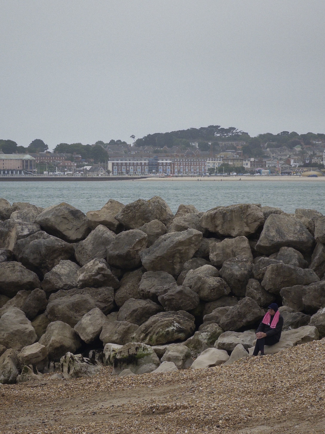 A lone person sits on pale rocks at the edge of a pebble beach, wearing dark clothing with a bright pink towel draped around their shoulders. A long line of large sea-defence boulders fills the foreground, while calm water and a distant waterfront town stretch across the horizon beneath an overcast sky.