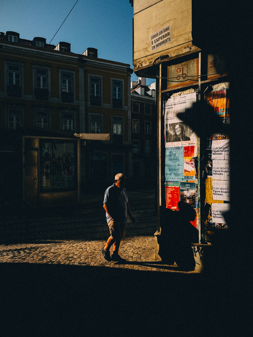 An older man walks along a cobblestone street in a sunlit urban area, passing a wall covered with colorful posters and flyers, with buildings in shadow behind him.