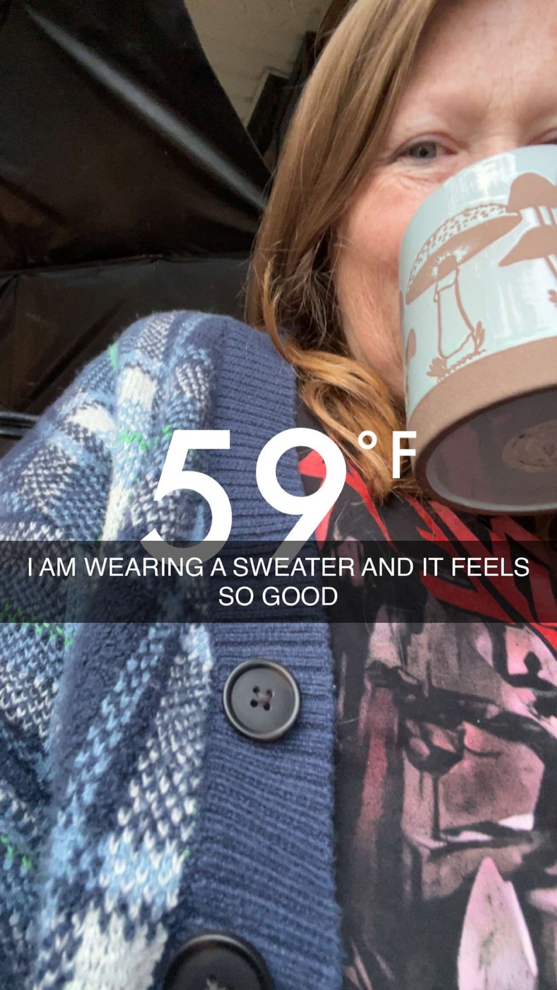 Snapchat image of a woman with freckles and blue eyes looking down at the camera, smiling behind her coffee cup, wearing a black tshirt with red letters (slayer) and a blue plaid cardigan sweater. Overlaid text says “59℉” and “I am wearing a sweater and it feels so good”