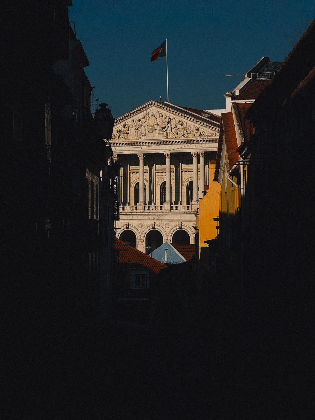Neoclassical building with ornate columns and sculptures, topped by a flag, framed by dark shadowed alleyways and colorful rooftops under a clear blue sky.