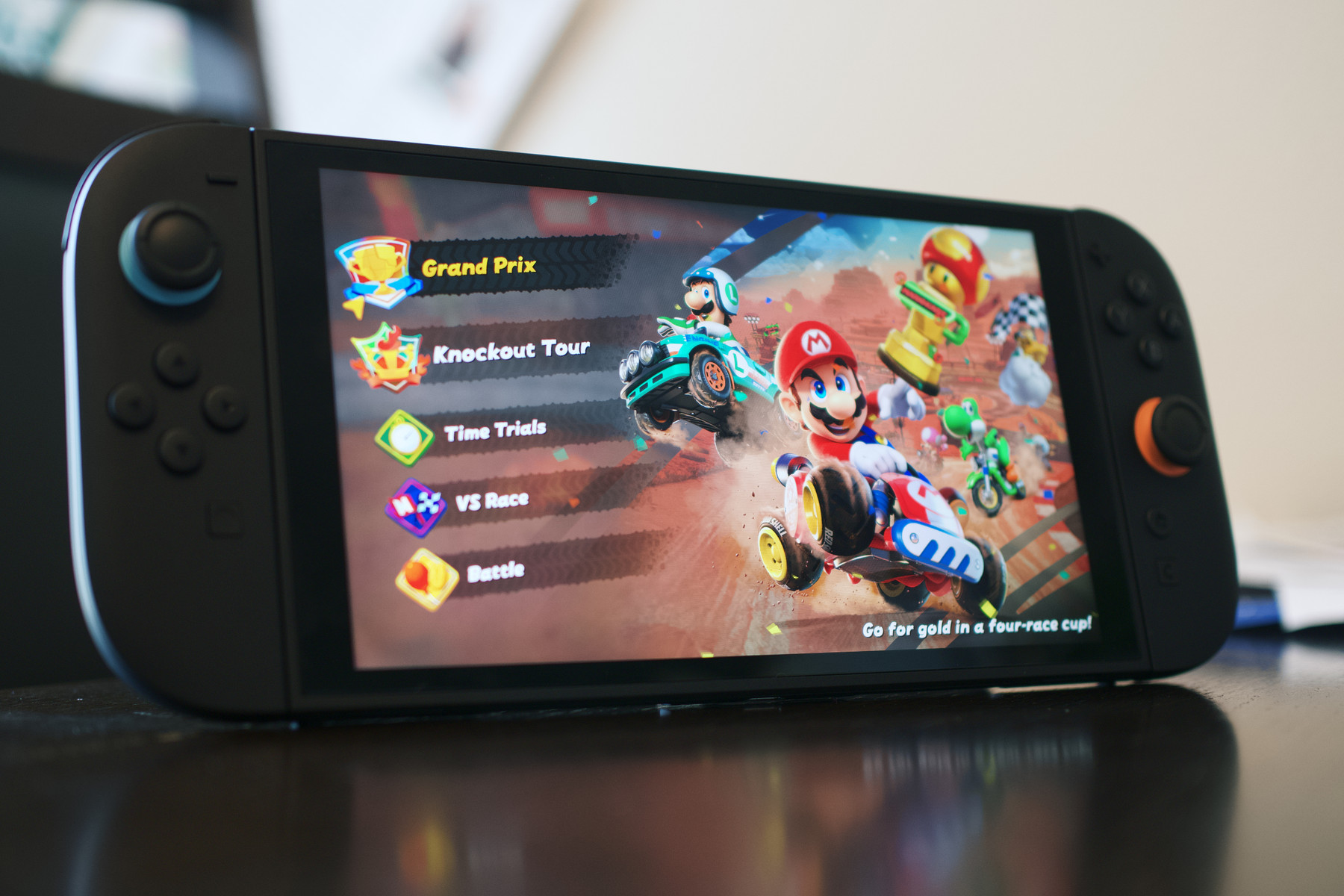 A handheld gaming console (resembling a Switch) displaying a colorful menu for a kart-racing game. The screen shows various modes like “Grand Prix,” “Knockout Tour,” “Time Trials,” “VS Race,” and “Battle.” In the background, characters like Mario, Luigi, and Yoshi are driving go-karts with items and course details visible around them.