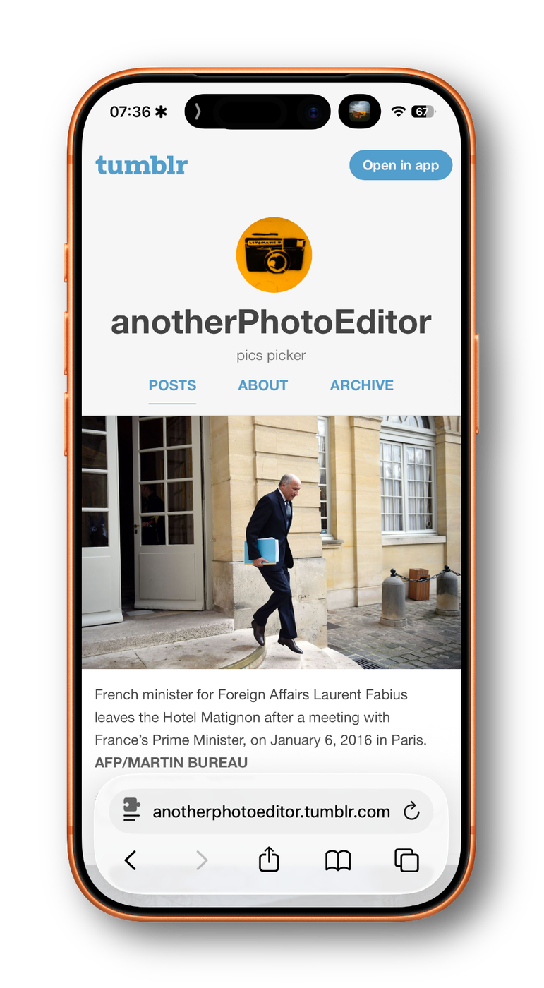 Smartphone displaying a Tumblr page for “anotherPhotoEditor” with a photo of a man in a suit carrying a blue folder, walking out of a building with tall windows and beige stone walls.