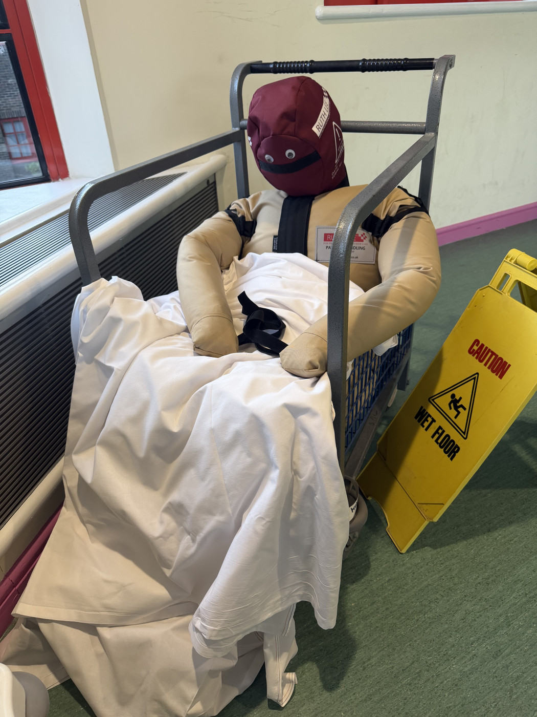 A mannequin dressed in a sits in a metal cage. Googly eyes have been fixed to its face.

The cage is covered with a white sheet, and a yellow caution sign reading "Wet Floor" is nearby.