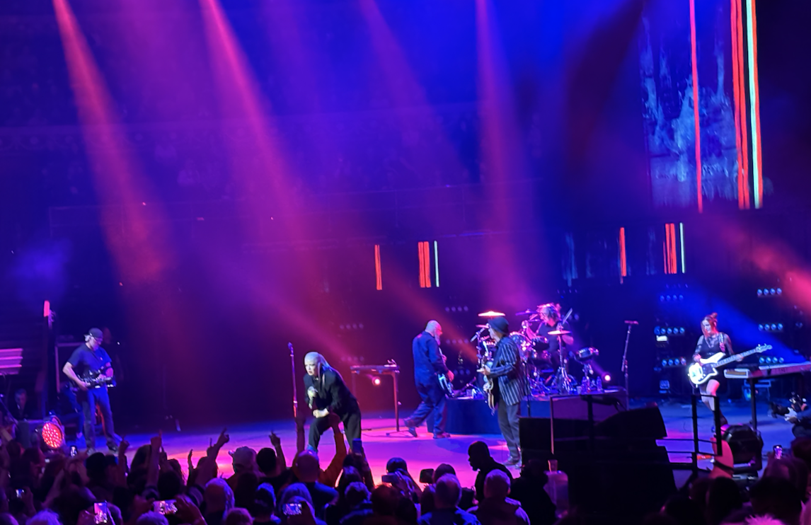 A band performs on stage under purple and blue lighting, with the lead singer at the front and musicians playing guitars and drums while an audience watches and raises their hands.