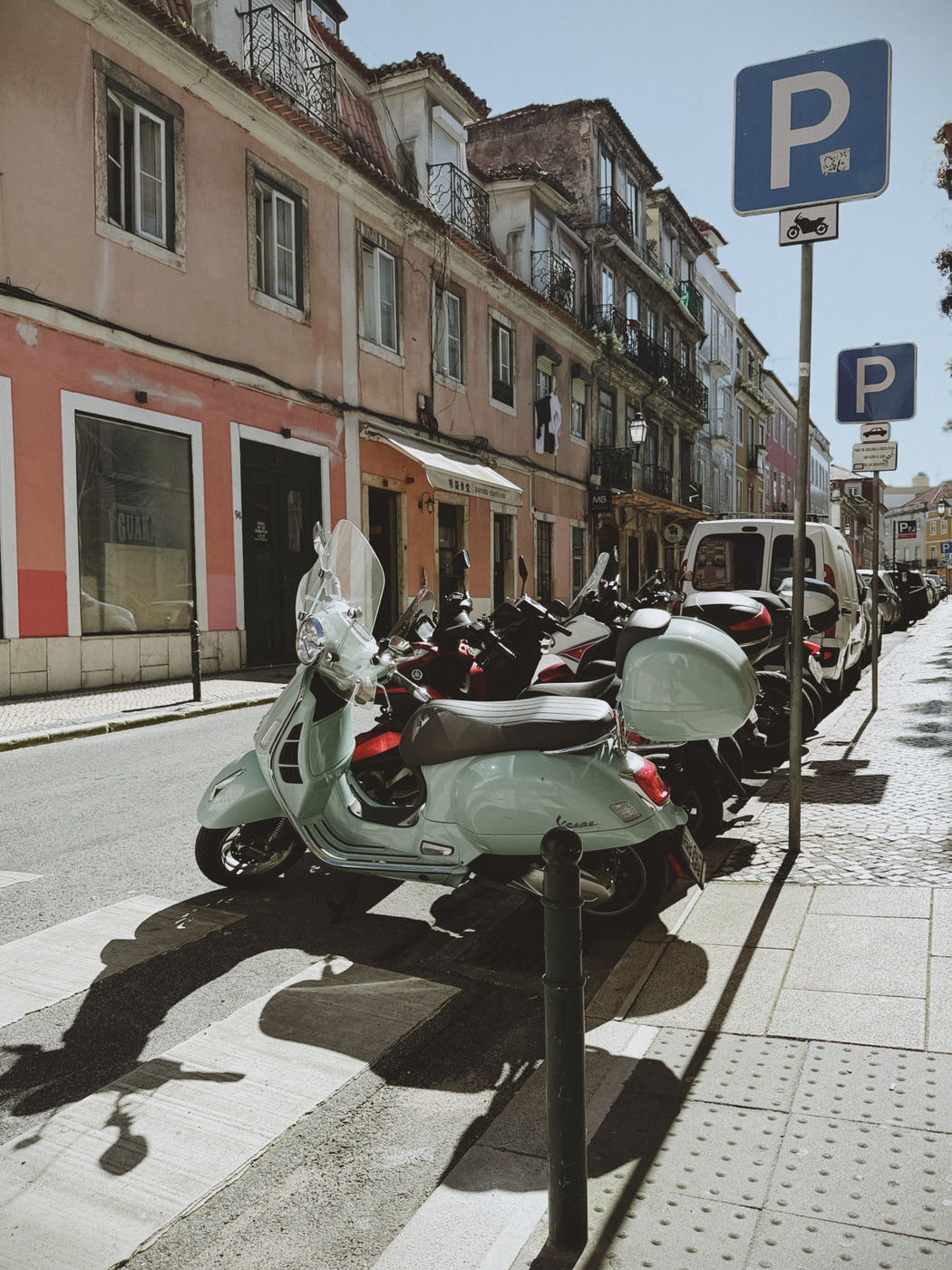 Row of parked scooters along a sunlit street with colorful old buildings and motorcycle parking signs.