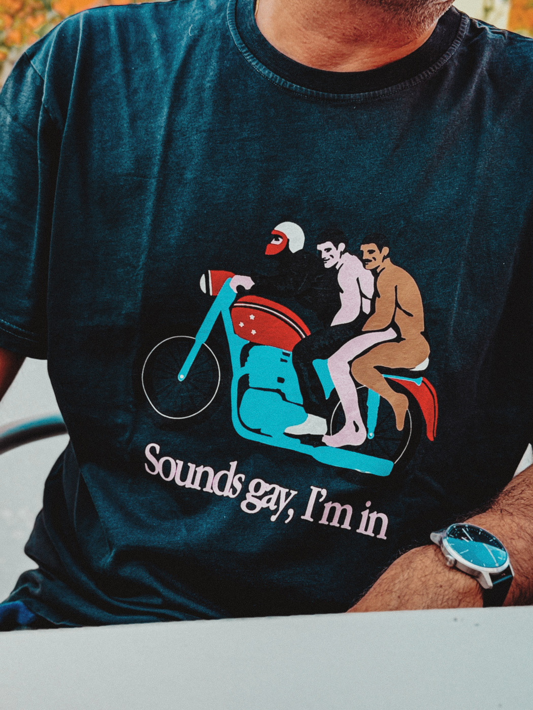 Person wearing a dark T-shirt with a colorful illustration of three men riding a motorcycle and the text “Sounds gay, I’m in.”