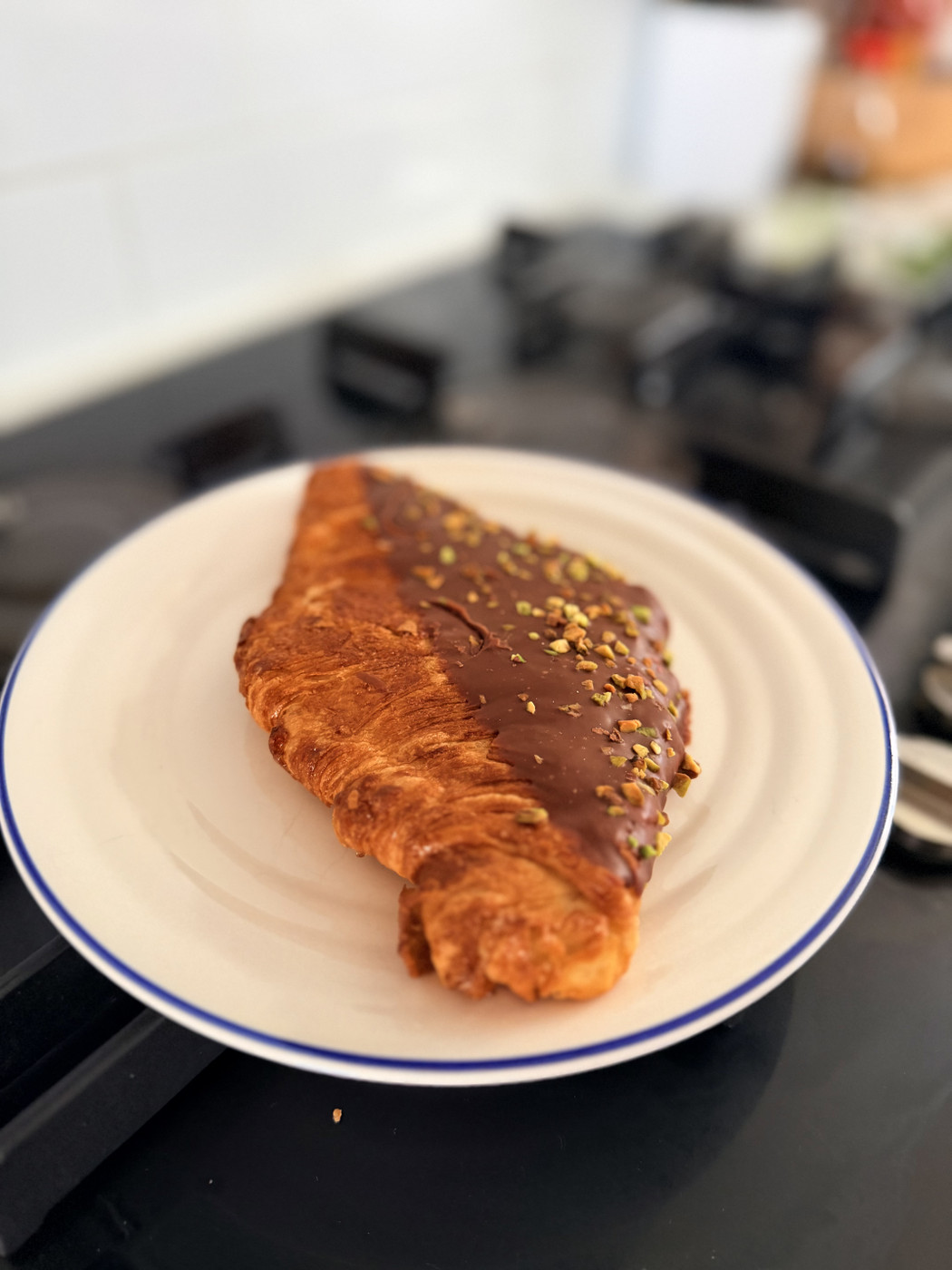 A freshly baked croissant with a glossy chocolate glaze and crushed pistachios sits on a white plate with a blue rim. The croissant is placed on a black stovetop, and the background is softly blurred, suggesting a cozy kitchen setting. The overall mood is warm and inviting, perfect for a delightful breakfast or snack.