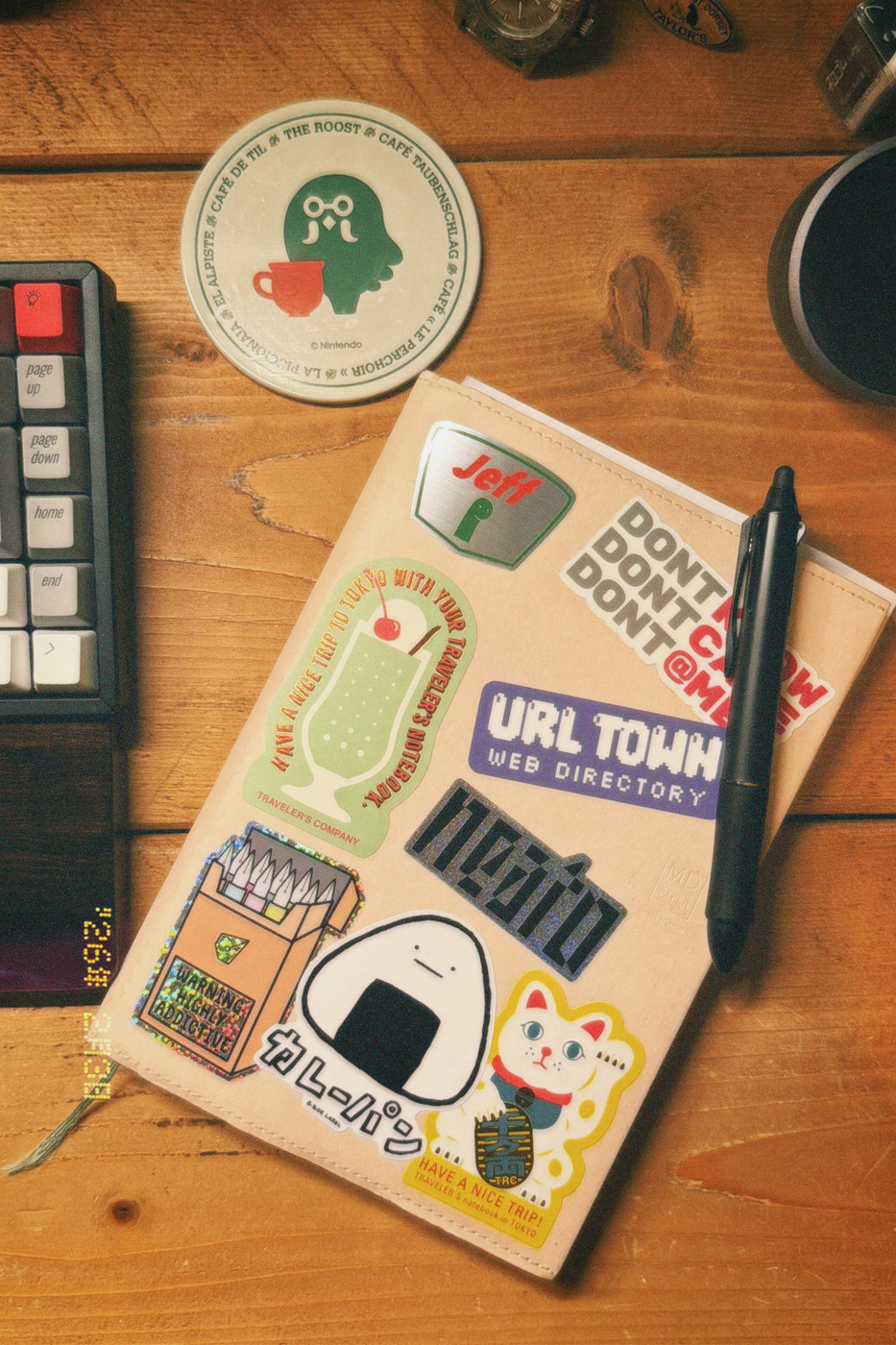 The image shows a neatly arranged desk scene photographed from above. At the centre is a light-coloured notebook or planner with stitched edges, covered in playful stickers—cartoon graphics, Japanese-themed icons, text labels, and retro-style designs. A black pen rests diagonally on top of the notebook.