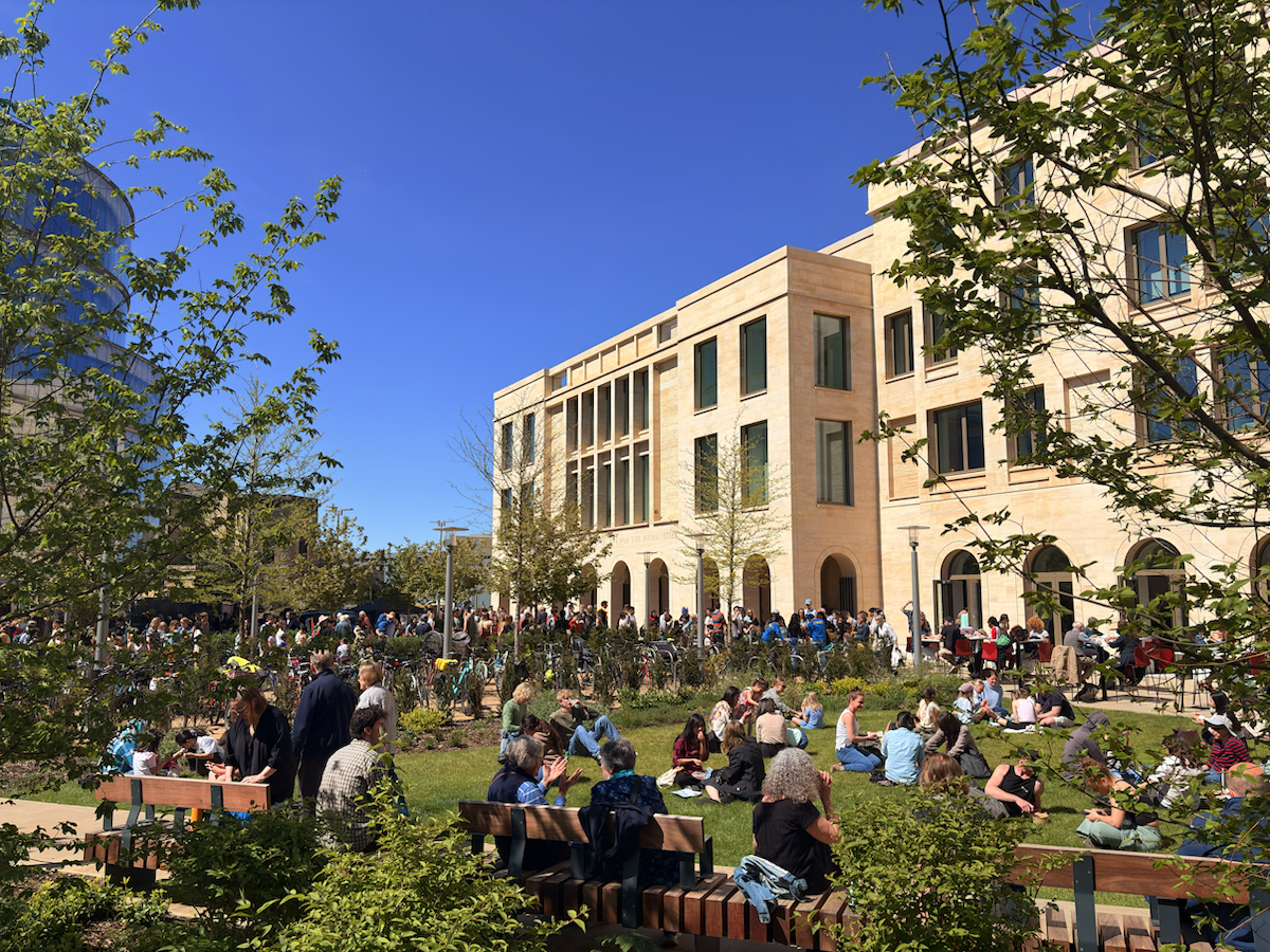 Crowd of people gathered on a sunny lawn outside a modern stone building with arched walkways, with groups sitting on the grass, others queuing near bicycle racks, and trees framing the scene against a clear blue sky.