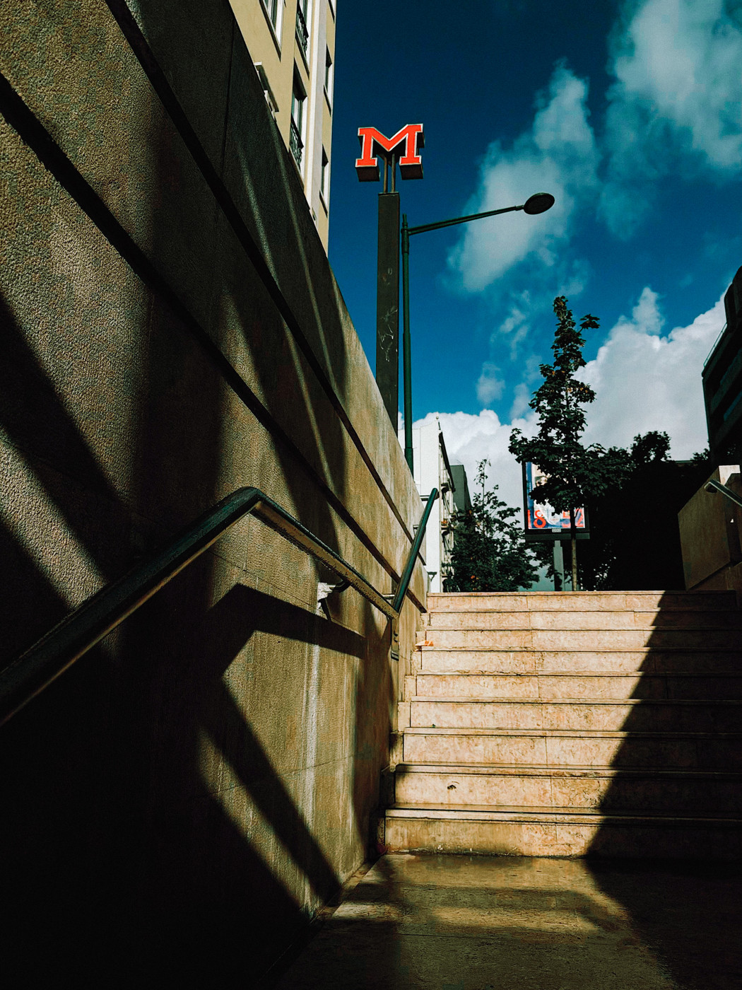 Stairway leading out of an underground area with sunlight casting shadows on the walls, a red “M” metro sign above, and a bright blue sky with clouds in the background.