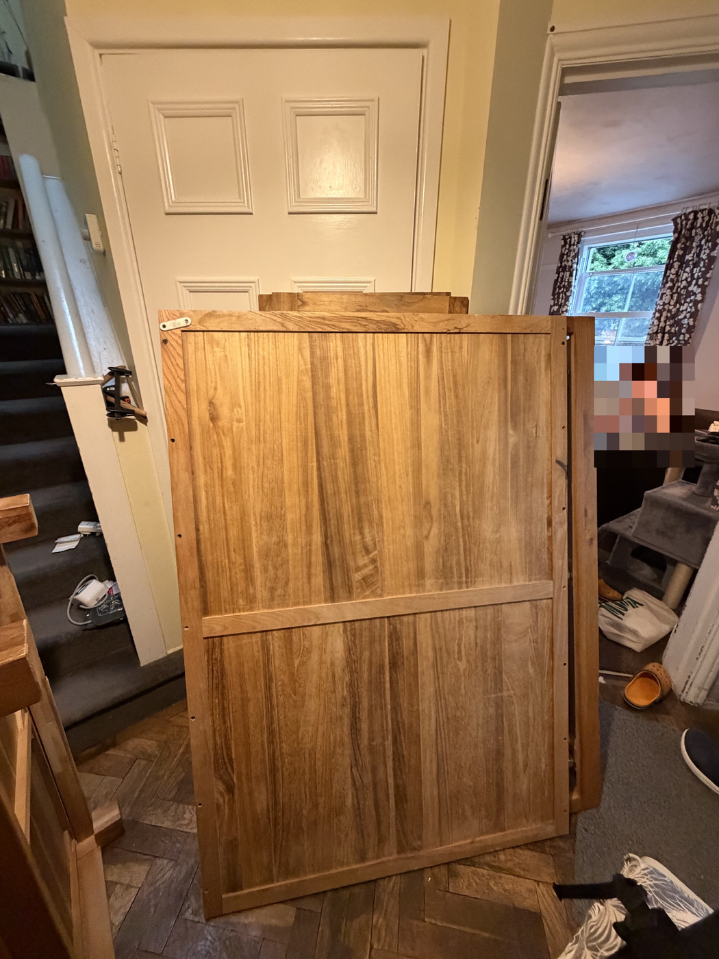 Large flat-pack wooden wardrobe panel leaning against a front door in a cluttered hallway with parquet flooring and stairs.