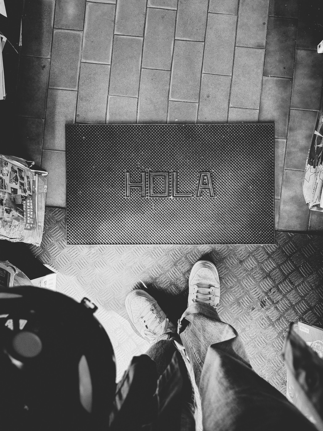 Black and white photo of a person standing on a metal-patterned floor in sneakers and jeans, facing a doormat with the word “HOLA” written on it, surrounded by scattered newspapers and magazines.
