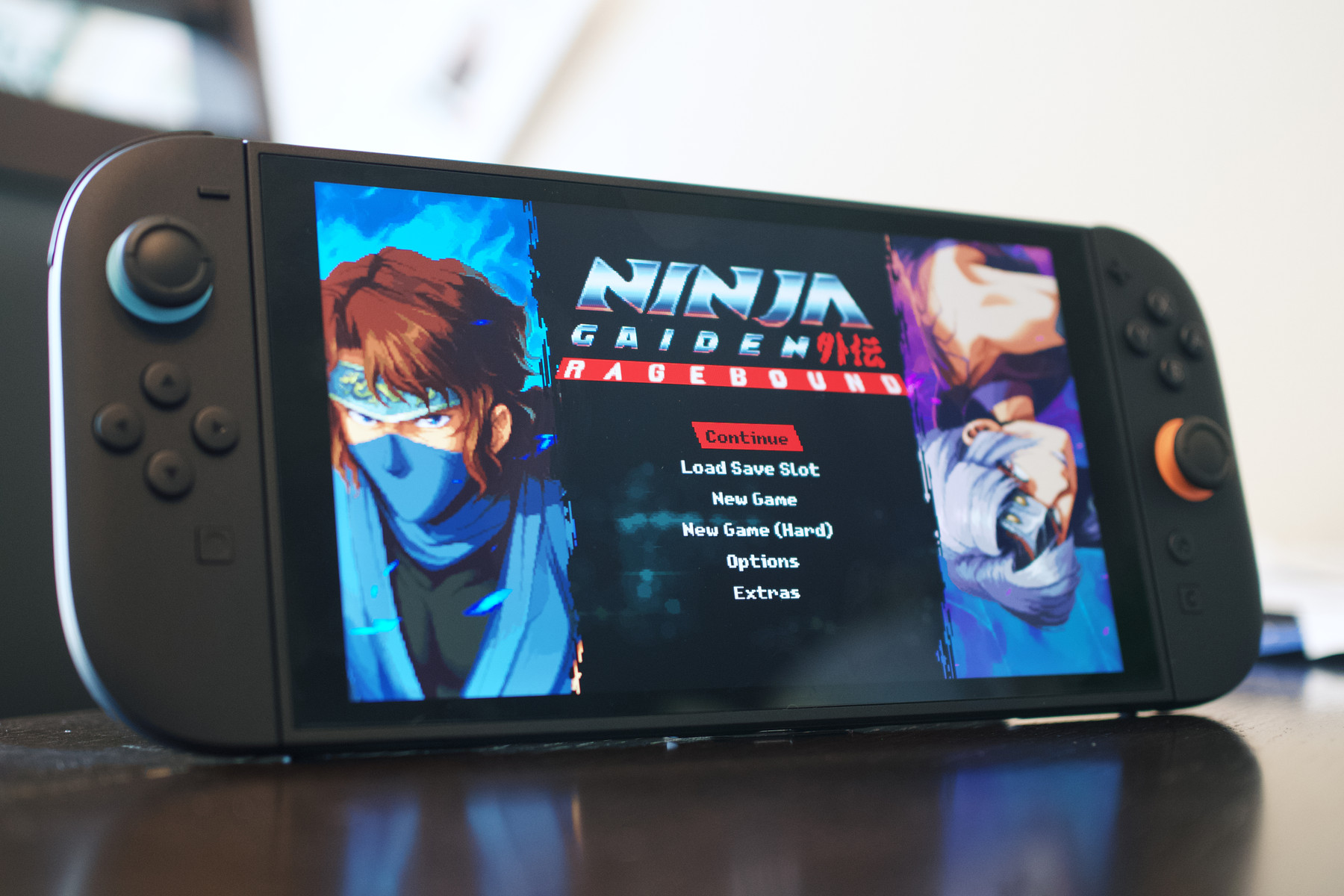 A handheld gaming console displaying the title screen for *Ninja Gaiden: Ragebound*. The screen shows a retro-styled menu with options like "Continue," "New Game," and "Options," flanked by anime-style portraits of two ninja characters. The console has black Joy-Cons with blue and orange joystick accents.
