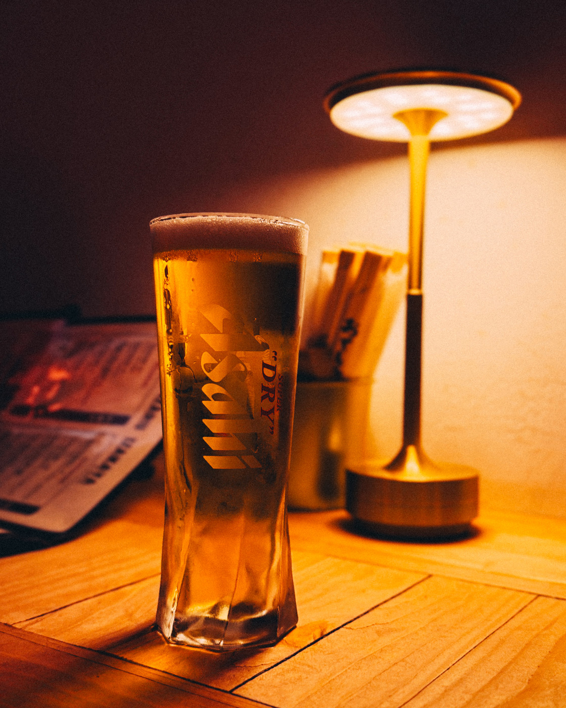 A tall glass of Asahi beer with a frothy head sits on a wooden table under warm ambient light from a nearby lamp, with a menu and chopsticks in the background.