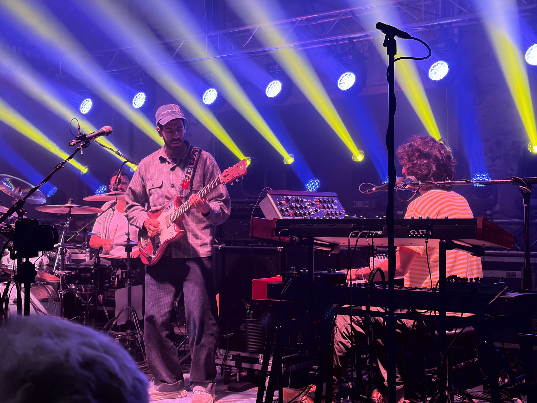 Three musicians on stage with colorful lights: a guitarist, a drummer, and a keyboardist, performing under a vibrant blue and yellow spotlight.