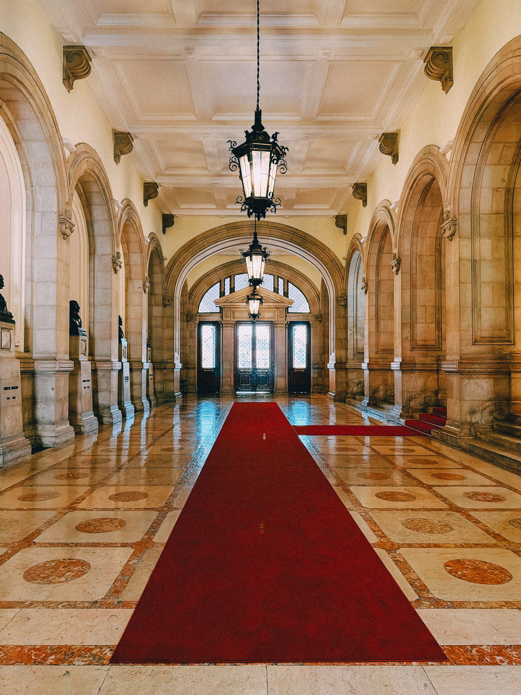 Grand hallway with arched doorways, marble floors, red carpet, and ornate hanging lanterns leading to double wooden doors with frosted glass panels.