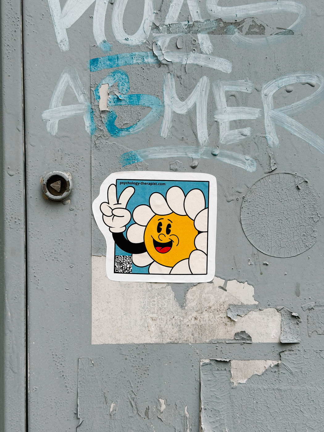 Sticker of a smiling cartoon flower giving a peace sign, placed on a weathered gray metal surface with peeling paint and graffiti.