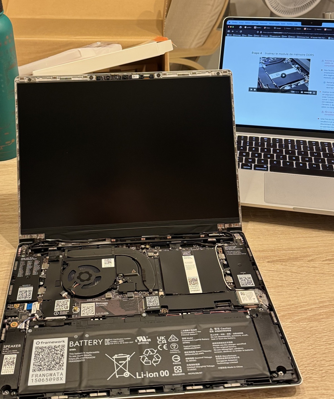 On the foreground, a Framework laptop rests on a table. The screen is black. It's open, we can see the internal components. In the background, another computer displays the Framework documentation with video guides.
