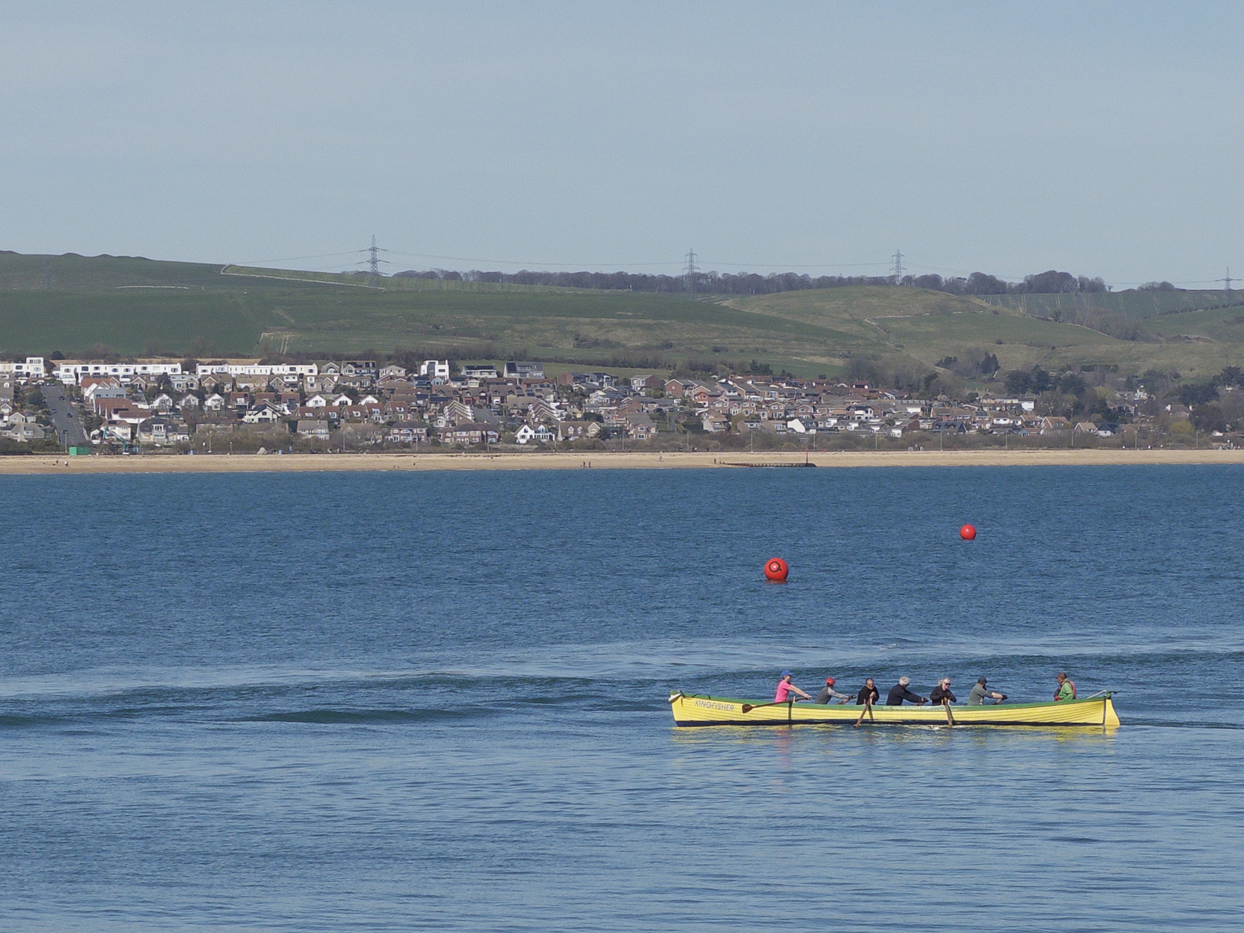 A rowing crew in a long yellow boat moving across open water, with a coastal town and green hillside in the distance.