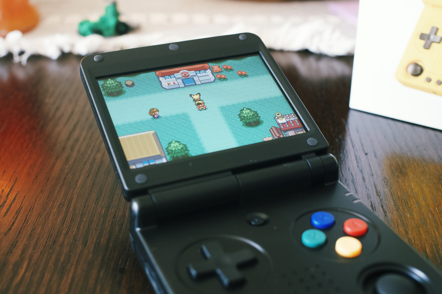 A black clamshell handheld gaming console displaying a Pokémon game on its screen, showing a character walking with Pikachu in front of a Pokémon Center. The console sits on a wooden table, with colorful buttons visible and a blurred yellow device box in the background.