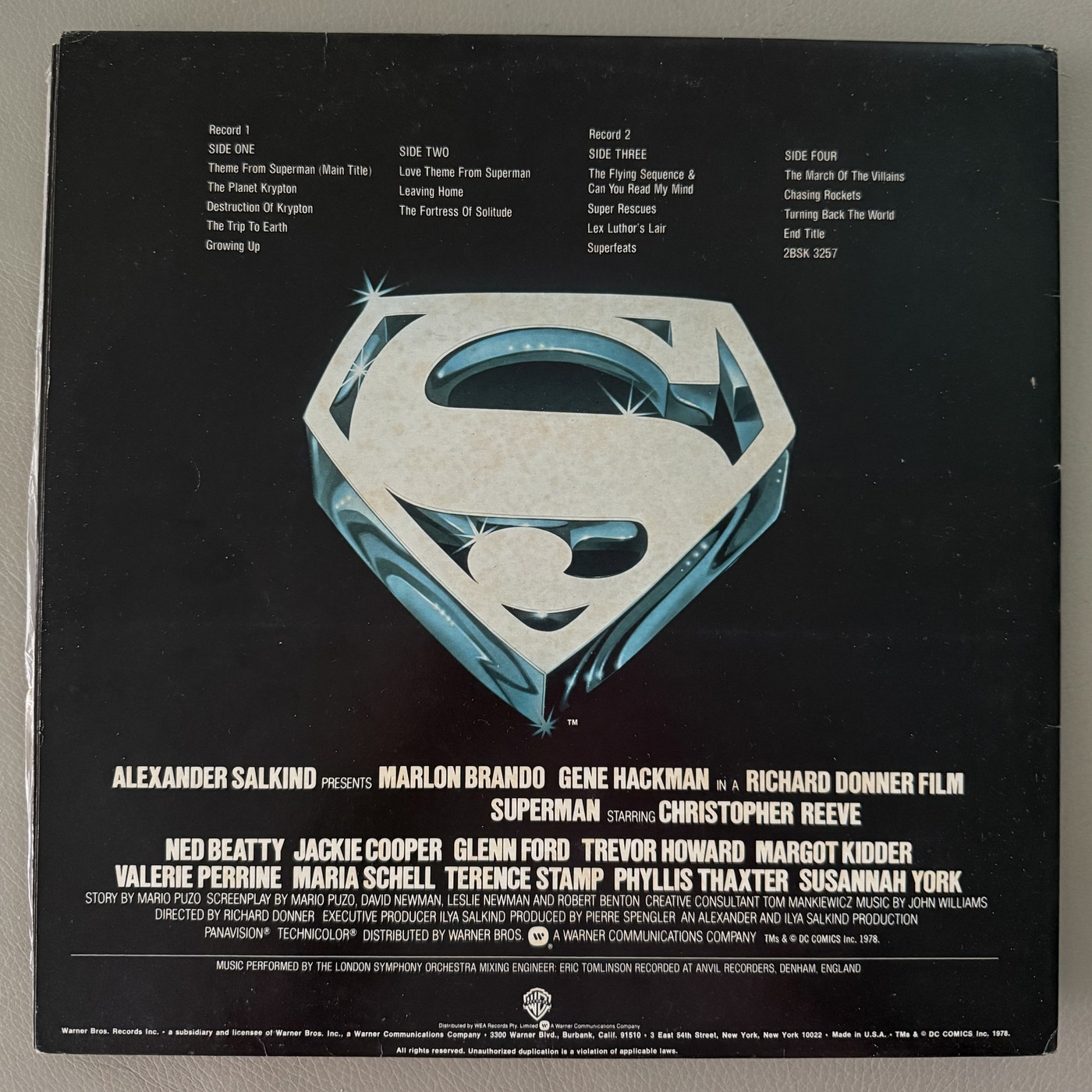 The back of the vinyl double album sound track for Superman The Movie. At the top it lists the songs on each side of the two records, has the large Superman S symbol in silvery blue in the middle, and underneath are the main credits for the movie.