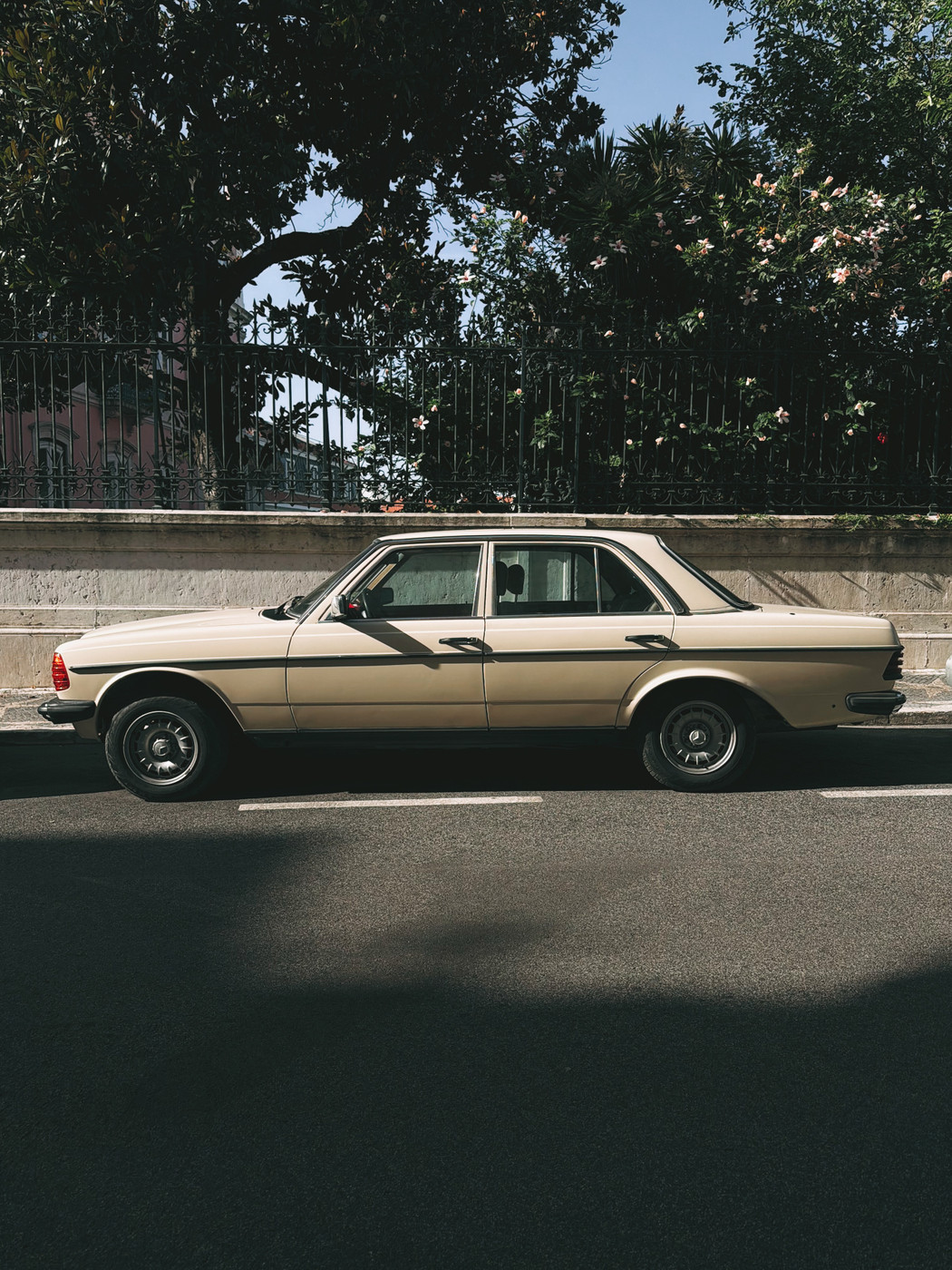 Vintage beige sedan parked on a quiet street with a wrought iron fence and lush trees in the background.