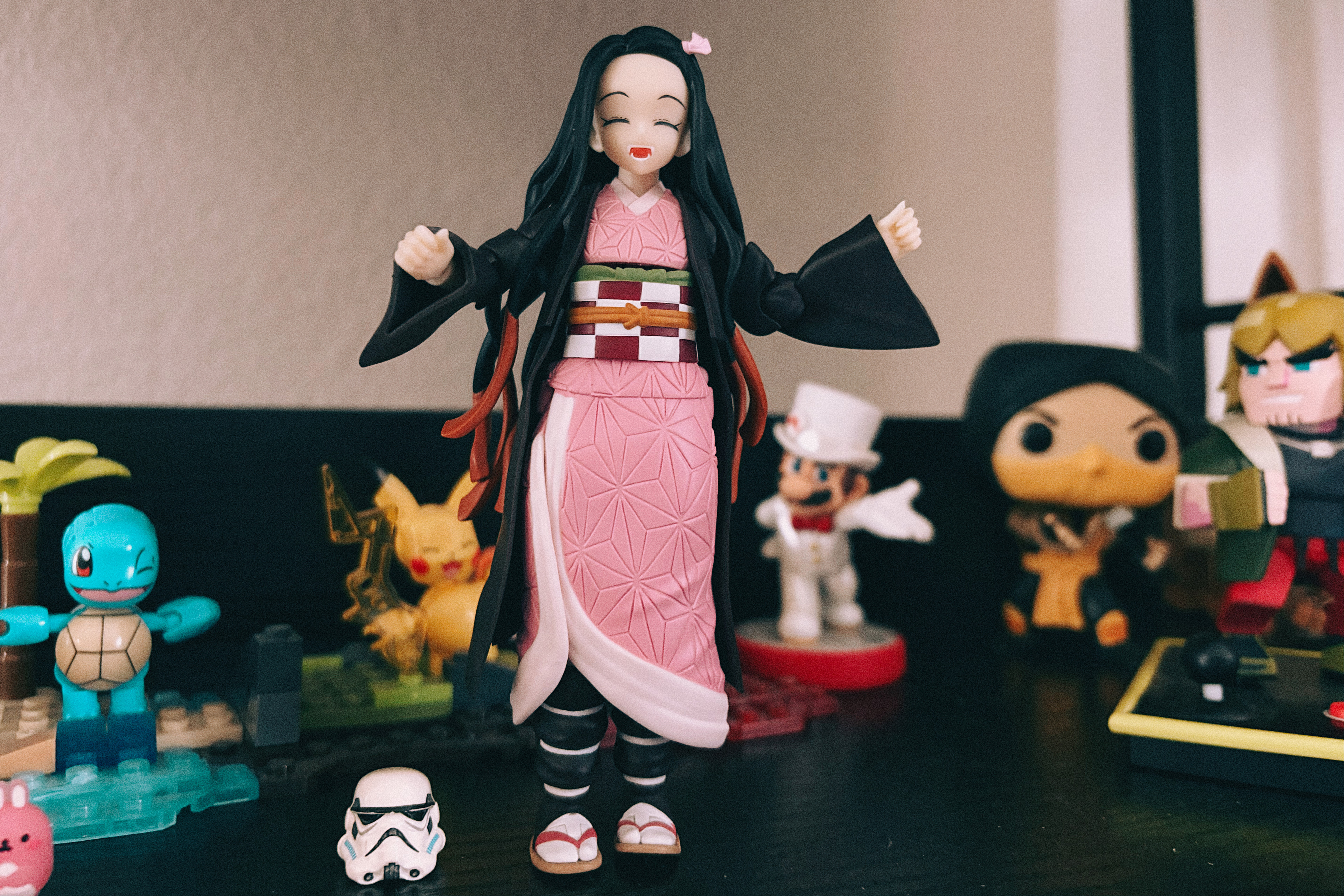 Uma estatueta de anime sorridente em um quimono rosa com longos cabelos pretos est&aacute; centralizada em uma prateleira, com os bra&ccedil;os ligeiramente levantados. Atr&aacute;s dele est&atilde;o v&aacute;rias figuras colecion&aacute;veis, incluindo um Squirtle e um Pikachu &agrave; esquerda e outros personagens estilizados &agrave; direita, com um pequeno capacete branco colocado na frente.