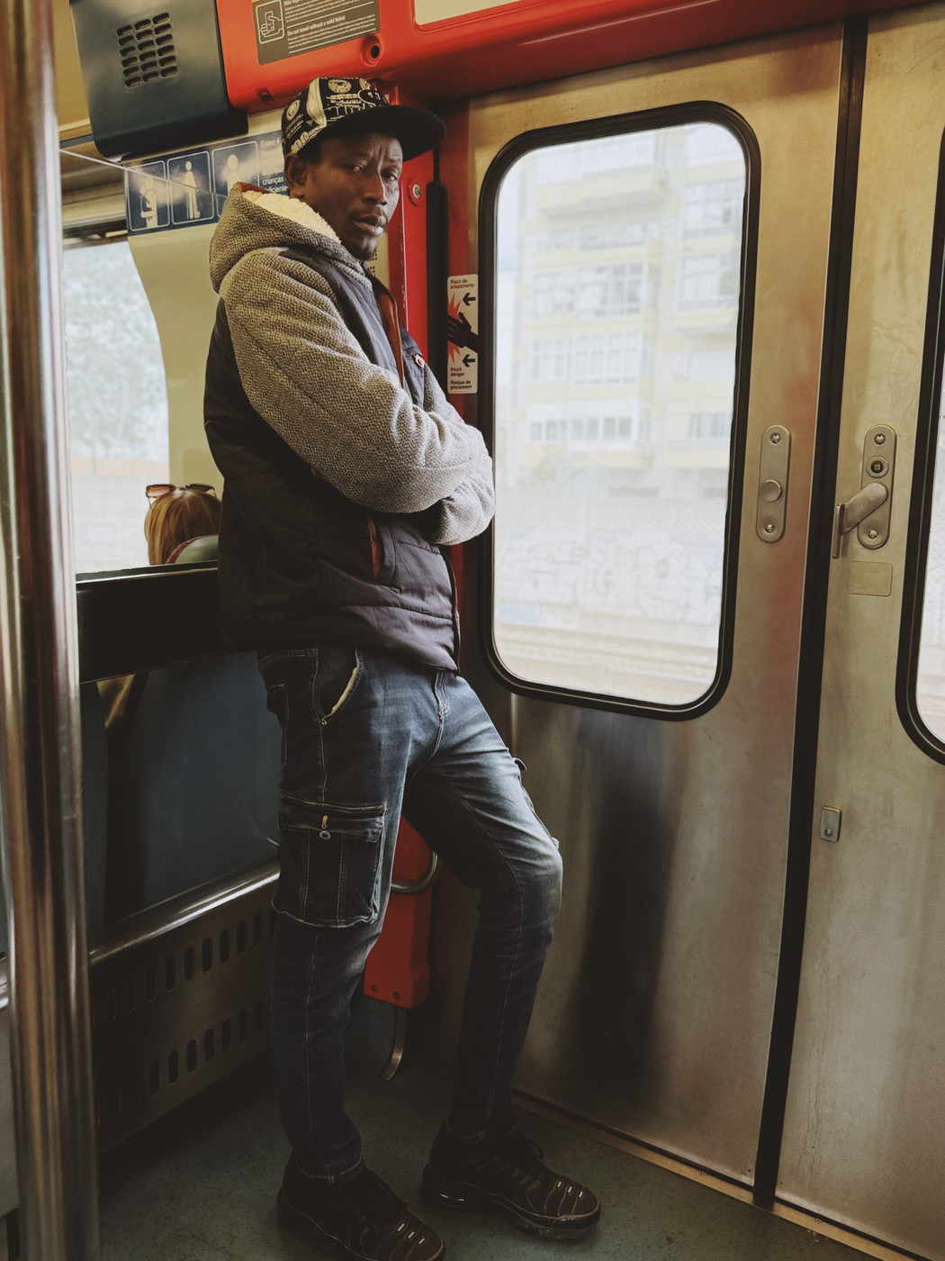 Man in a gray hoodie, black vest, jeans, and a cap leaning against a red and silver train door with arms crossed, while another passenger sits nearby looking out the window.