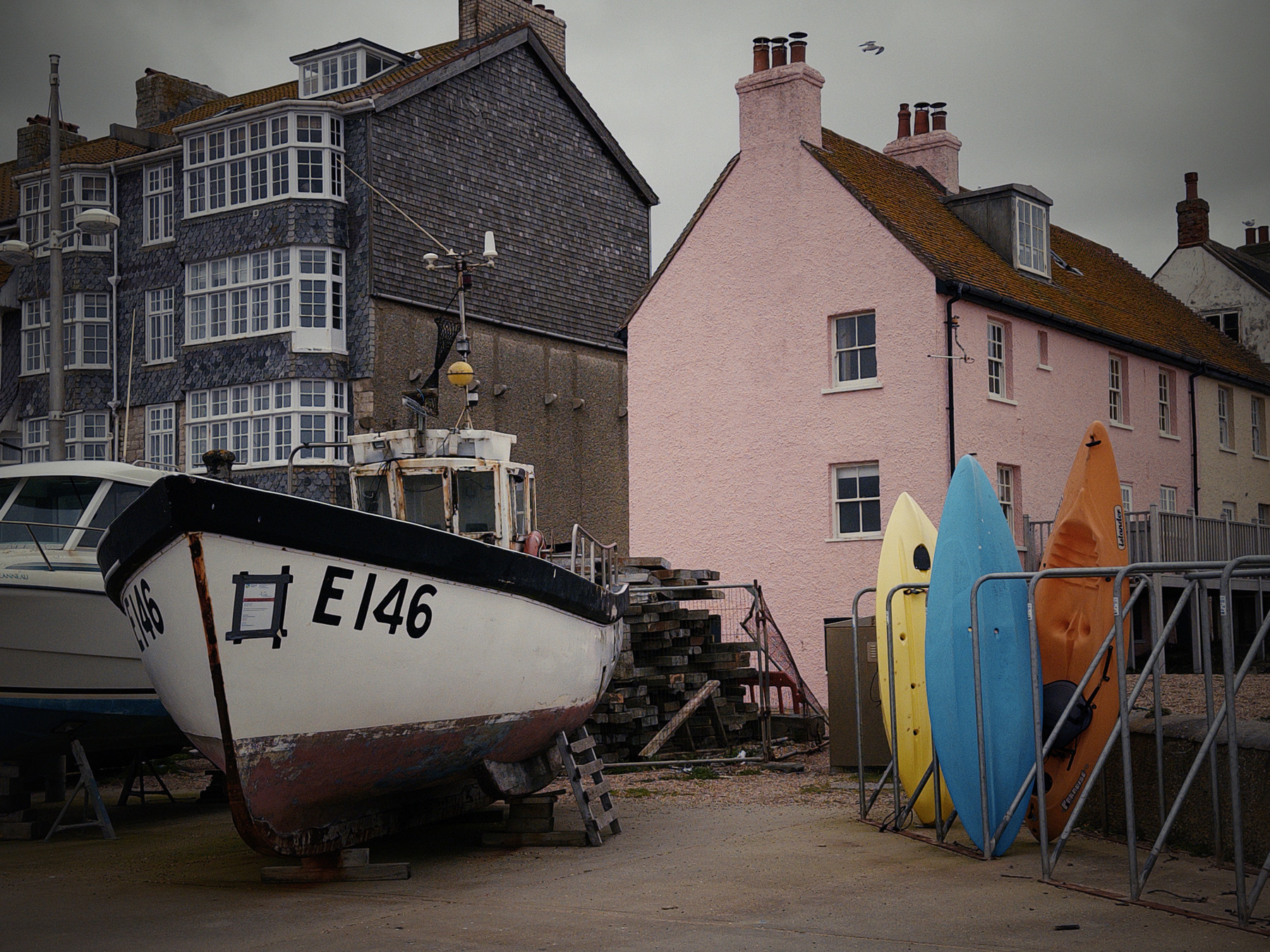 A fishing boat on dry land next to multi-coloured buildings under a grey sky. The mood is calm and quiet.
