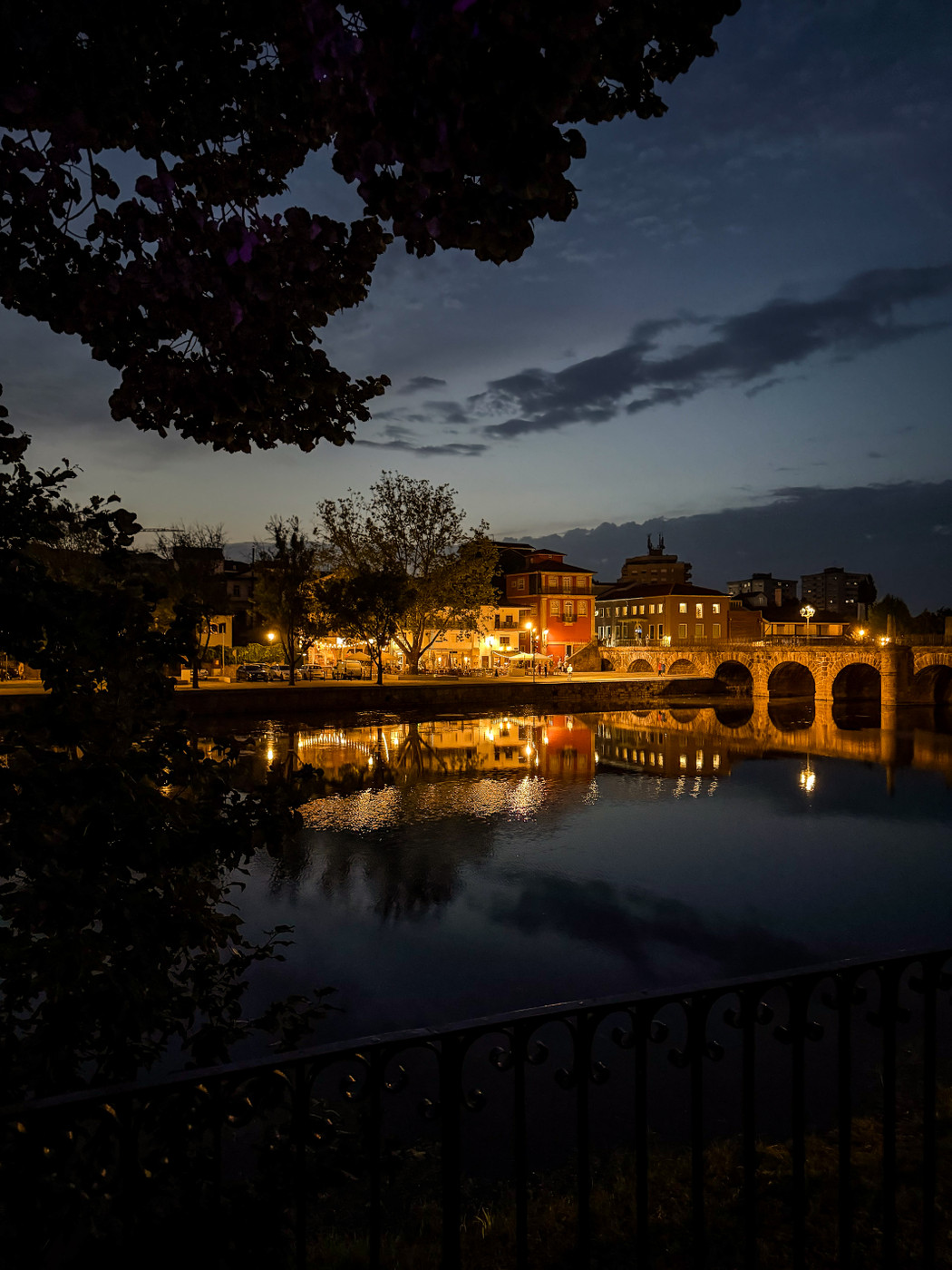Nighttime view of a riverside town with illuminated buildings reflecting on the calm water, a stone bridge with arches, and tree branches framing the scene.