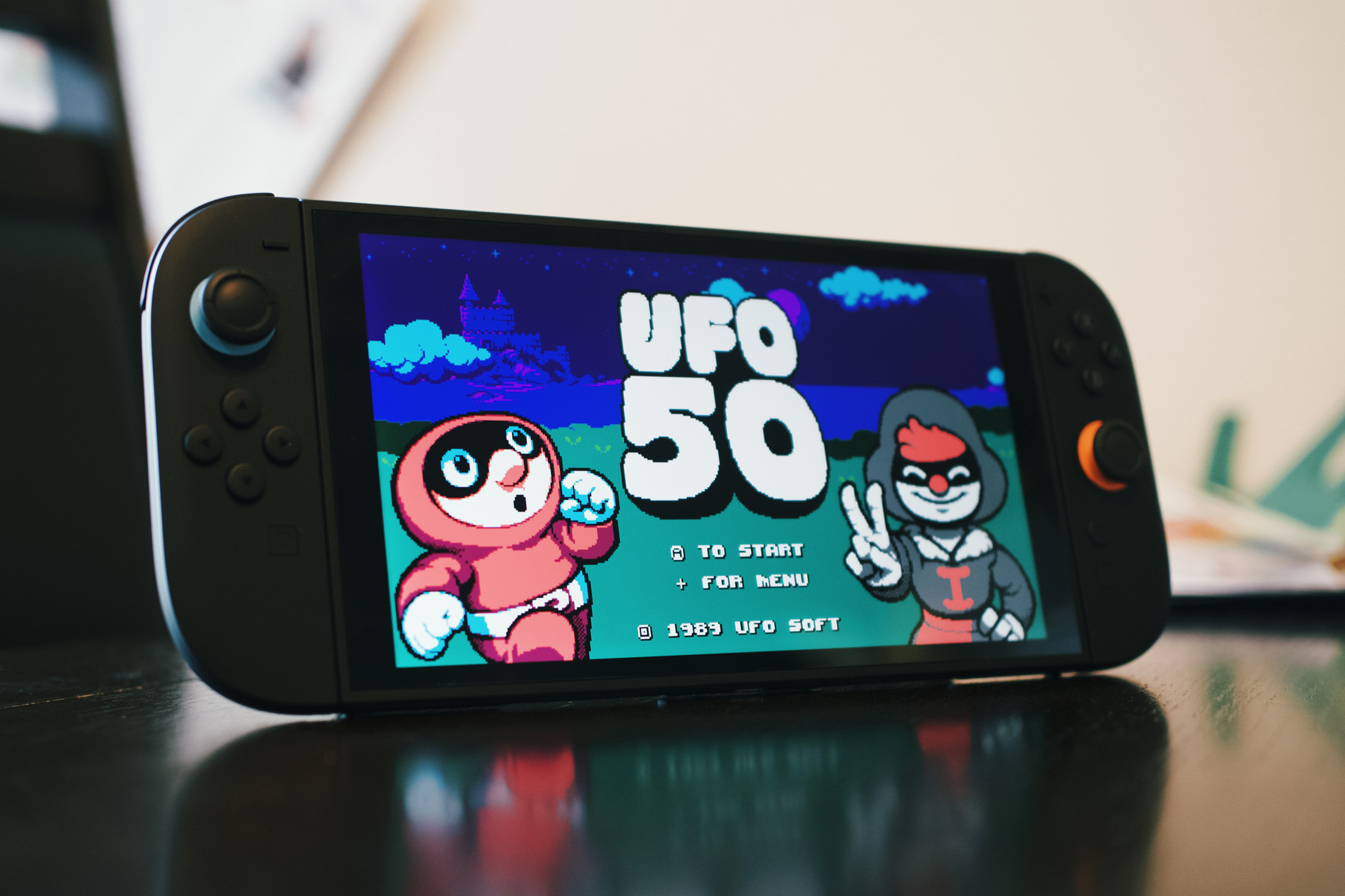A Nintendo Switch console on a table displaying the retro-style game UFO 50. The screen shows colorful pixel art of two cartoon characters: one in a red suit with white gloves and another in a hoodie making a peace sign. The game’s title “UFO 50” appears in large white letters, with menu options below.