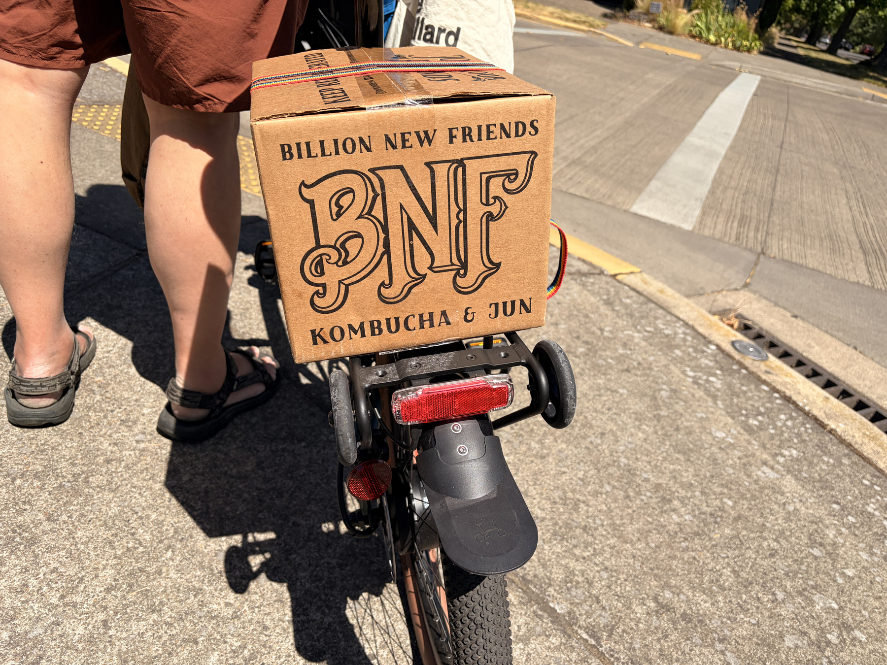 A cardboard box that says BNF Kombucha and Jun on the back of a Brompton bike, being walked by a devastatingly handsom man, whose glorious sculpted calves are the only parts of him visible.