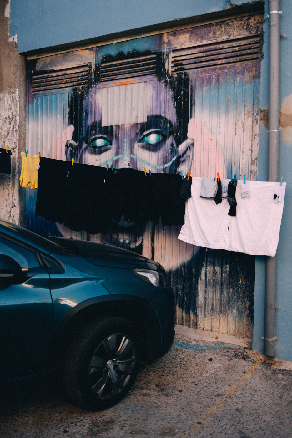 Street art of a face with glowing eyes on a weathered garage door, partially obscured by hanging laundry and a parked blue car.