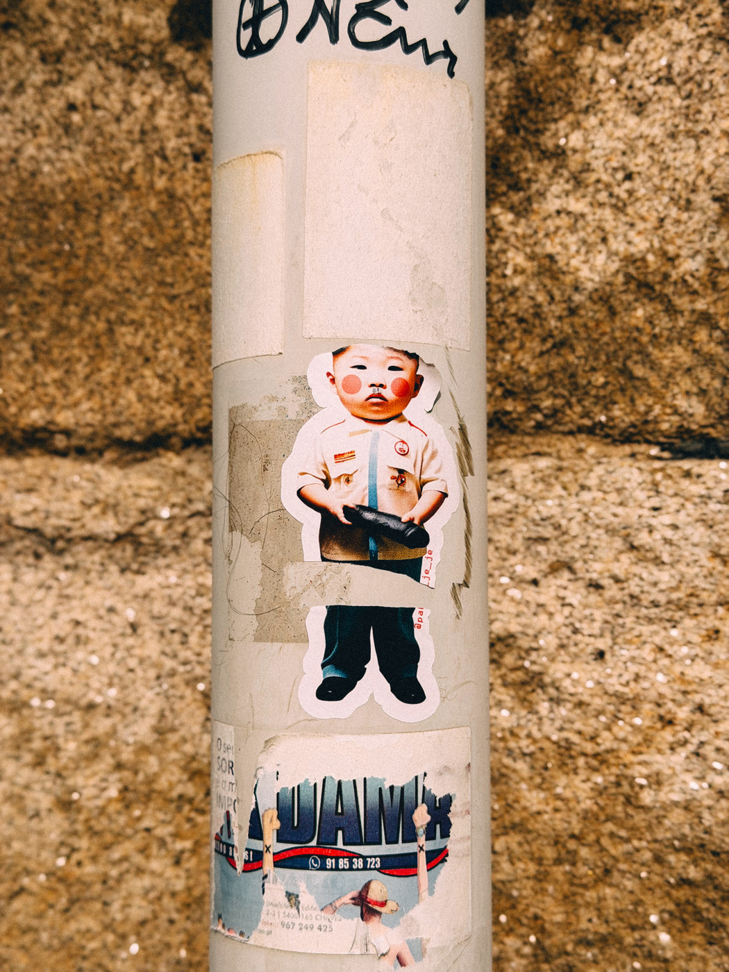 Sticker-covered pole with a central sticker of a child in a uniform holding an object, surrounded by worn and partially torn labels against a stone wall background.