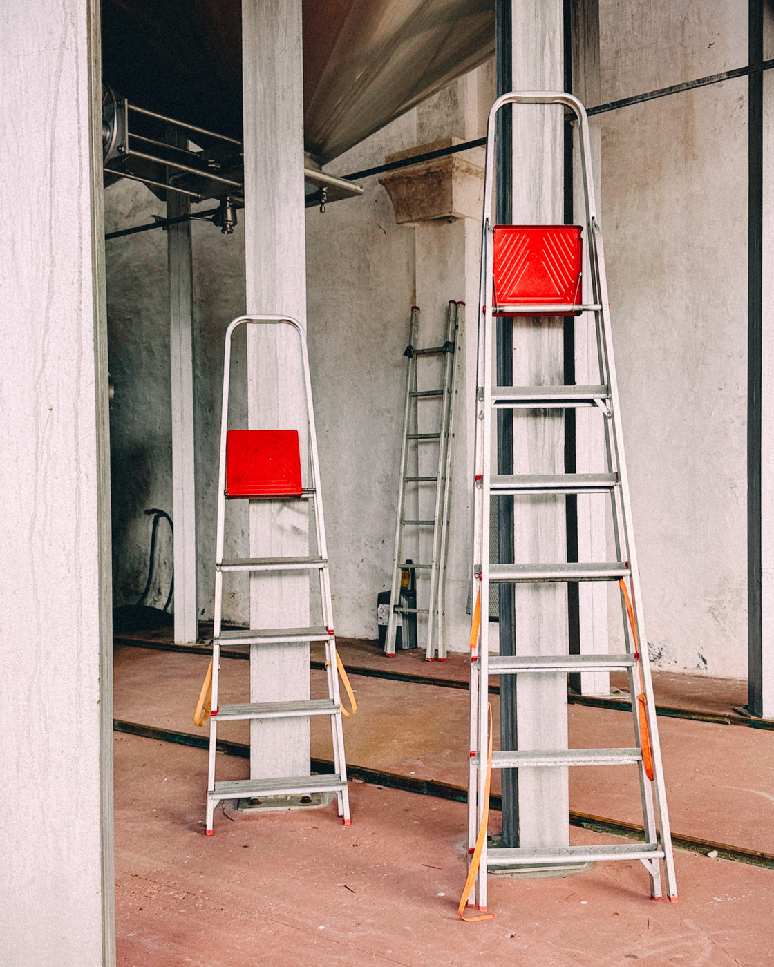 Two aluminum step ladders with red tops leaning against columns , while a third ladder leans against the back wall.