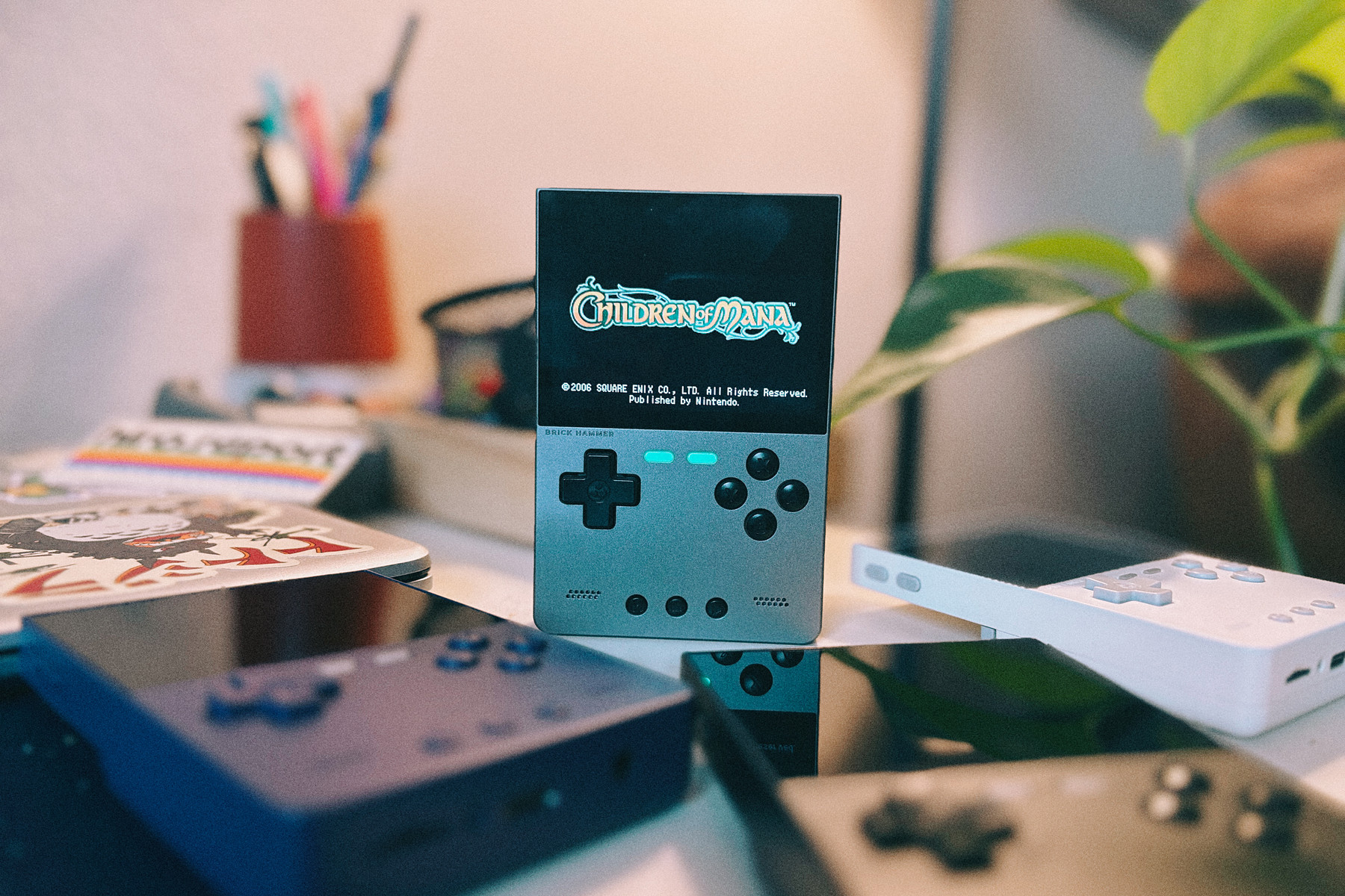 A small retro handheld game console on a desk, showing the “Children of Mana” title screen, with other game controllers blurred in the foreground and a plant in the background.