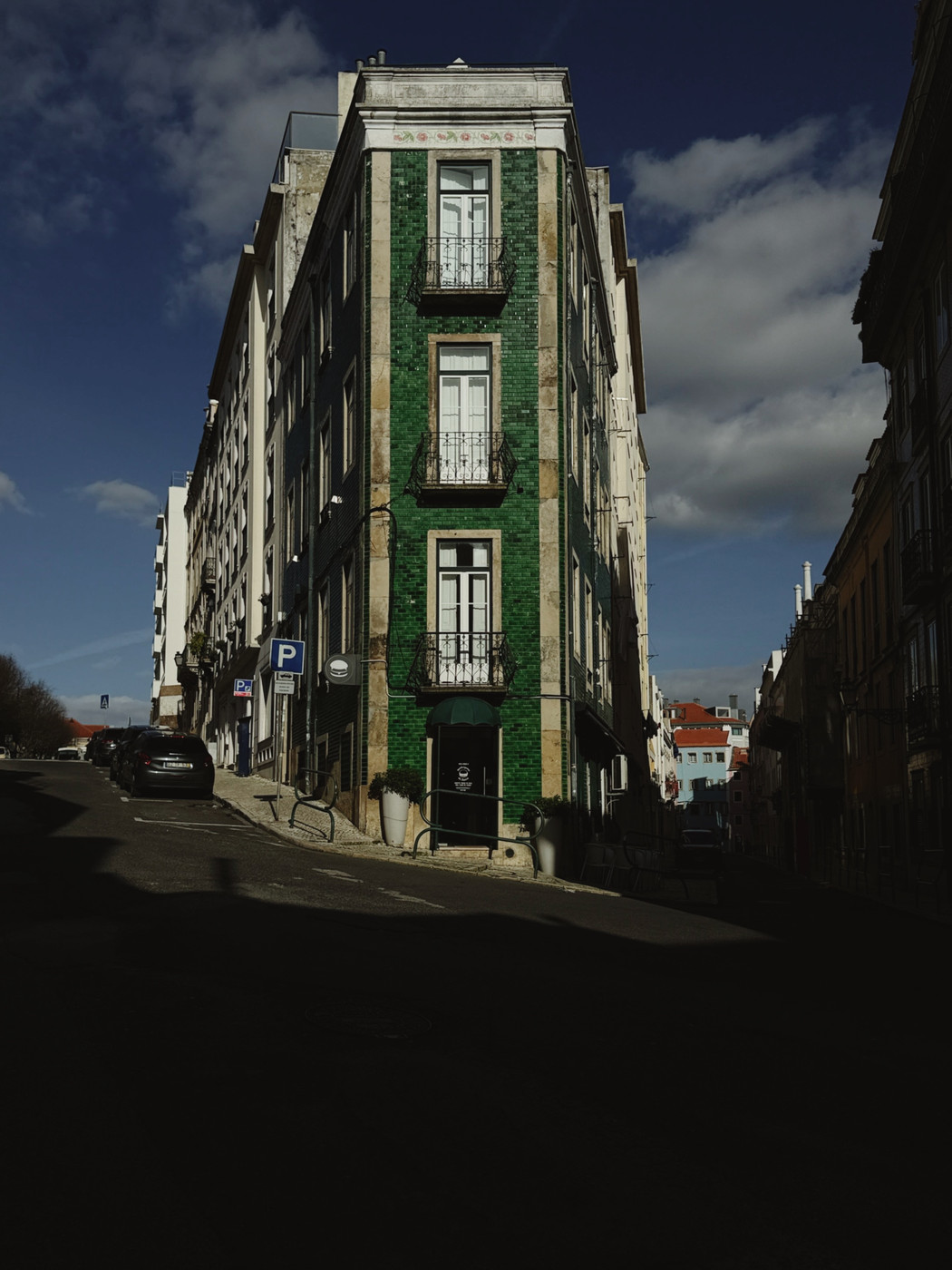 Narrow green-tiled building on a sunlit street corner with balconies, surrounded by shadows and a blue sky with scattered clouds.