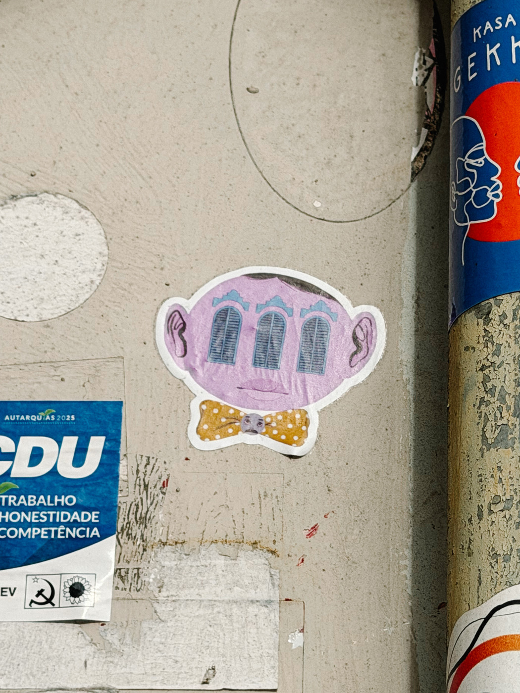 Sticker of a purple cartoon face with three blue windows for eyes, ears on the sides, and a yellow polka-dotted bow tie, placed on a worn wall with other posters and stickers.
