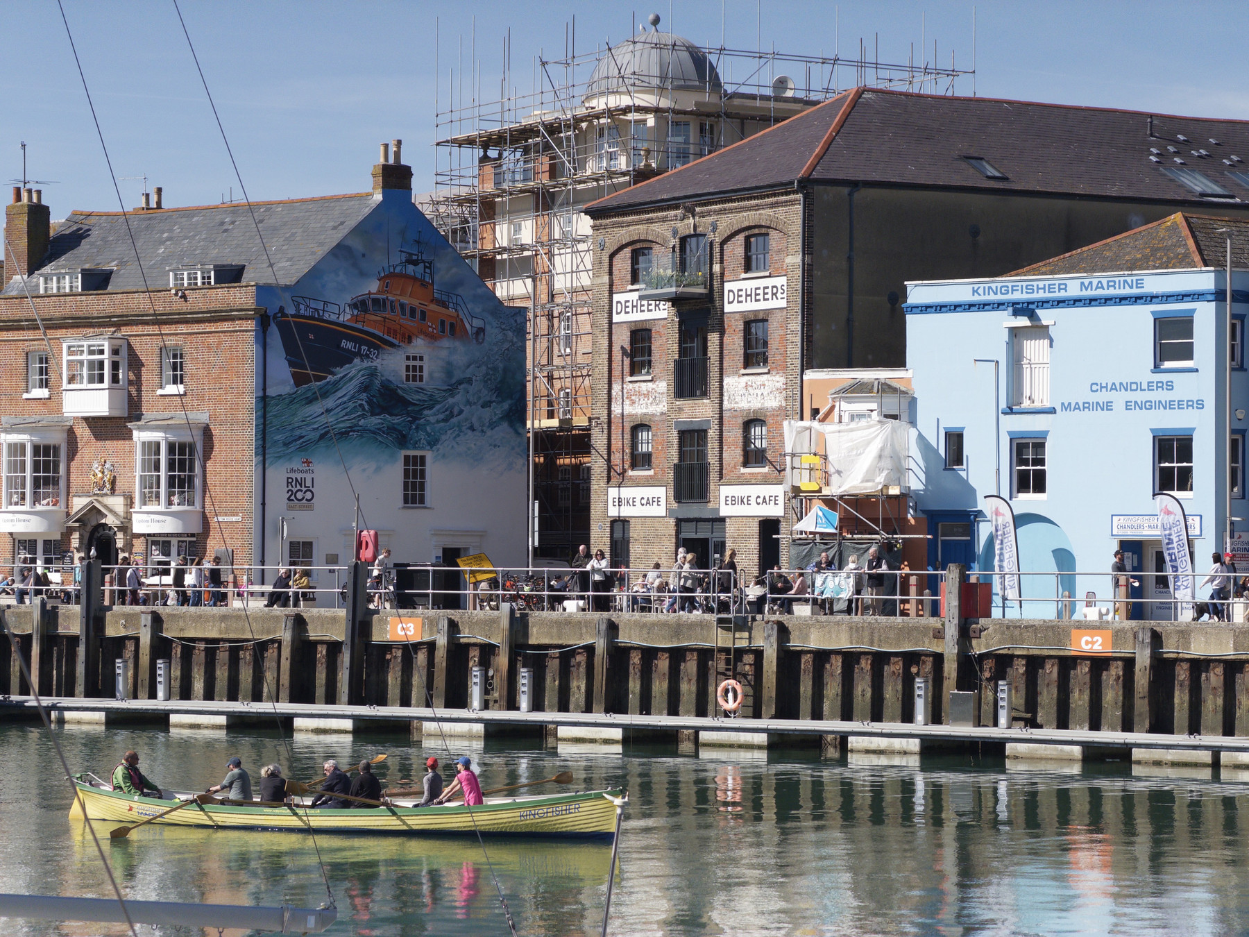 A long rowing boat with a team paddling along a harbour, with brick buildings and a large mural visible on the waterfront behind them.