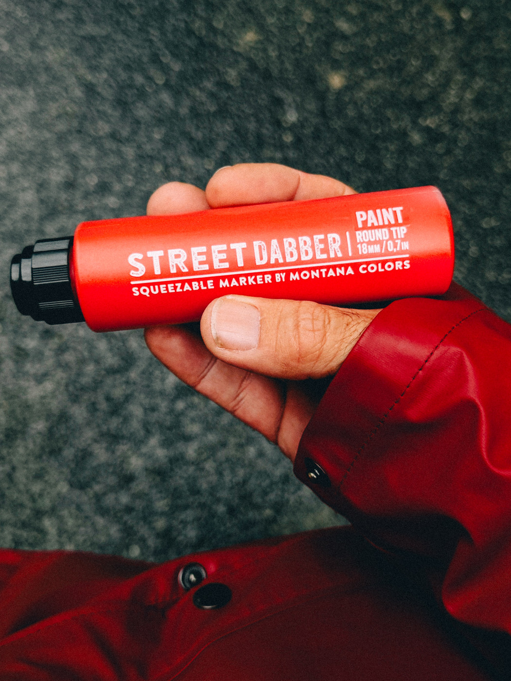 Hand holding a red Montana Colors Street Dabber paint marker with a round tip against a textured gray background.