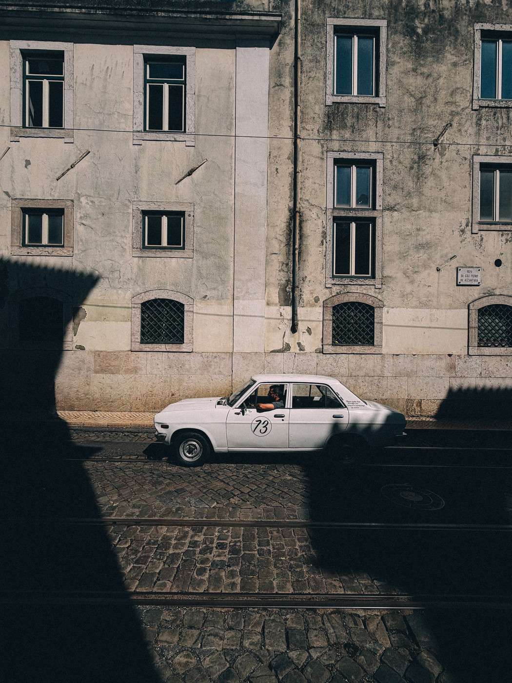A vintage white car with the number 73 on its door drives along a cobblestone street beside an old, weathered building with multiple windows and strong shadows cast across the scene.