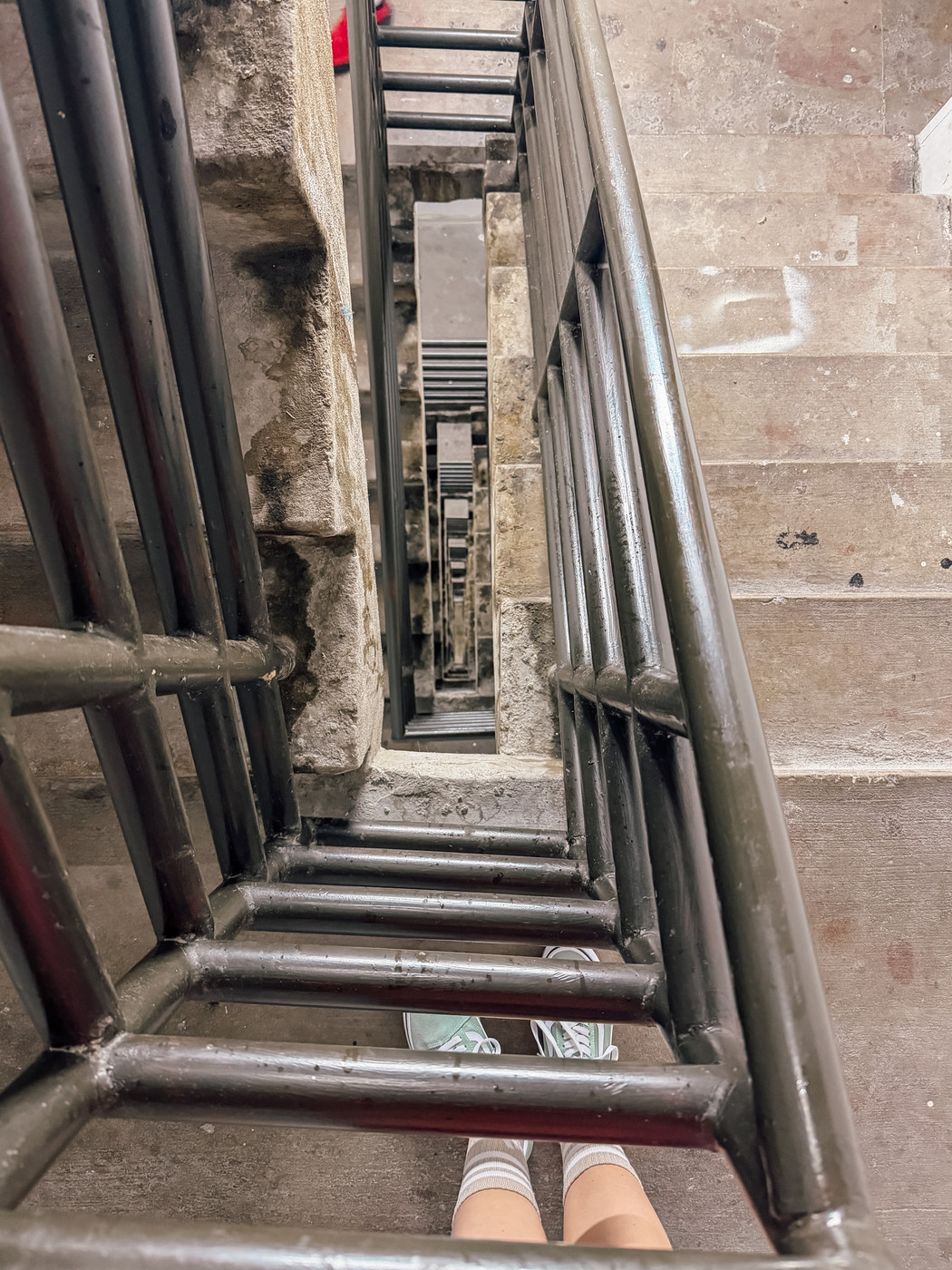 A staircase with multiple flights is viewed from above, showing a dizzying downward perspective and a person wearing sneakers.