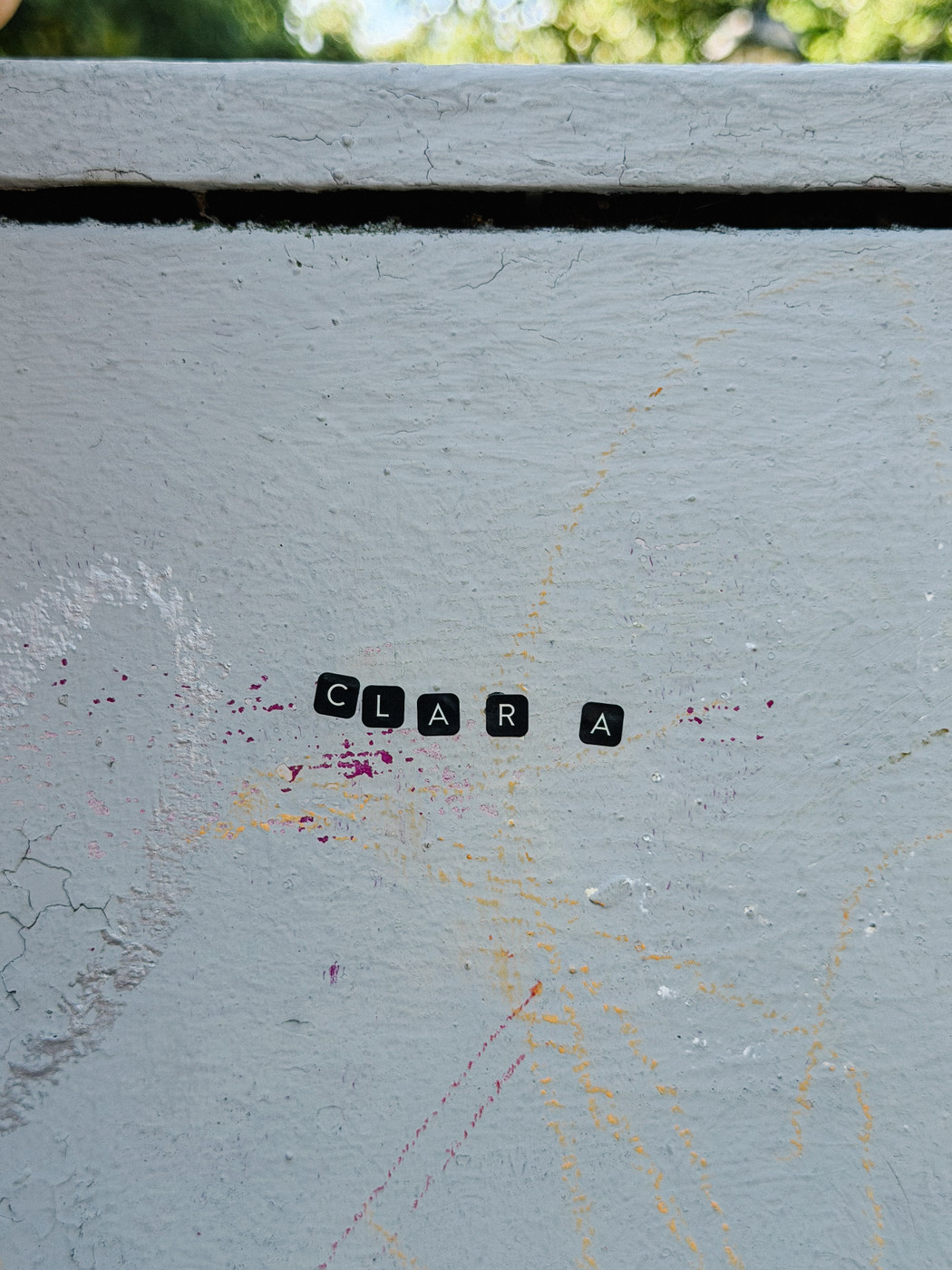 Black letter stickers spelling “CLARA” on a light gray utilities box with faint colorful scribbles.