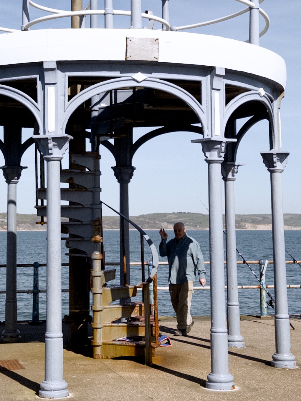 Man walking near a spiral staircase structure on a pier, with fishing rods and the sea in the background.